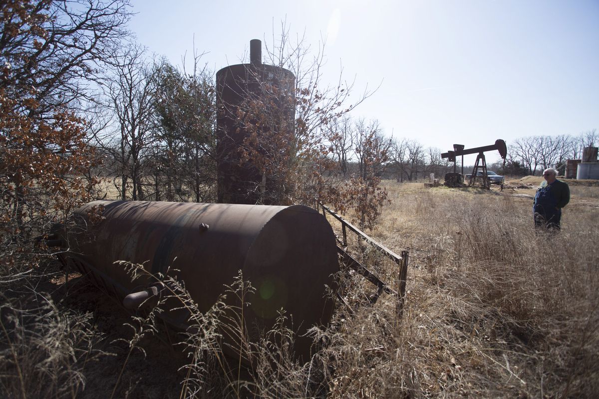An older man stands looking at weed-covered oil field equipment. An oil pump is in the background.
