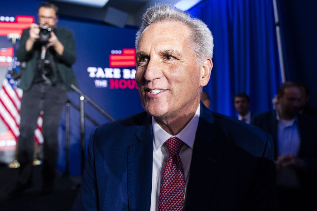 McCarthy, clean shaven in a navy suit, red tie, and white shirt, smiles. His silver hair shines under a bright stage light. Behind him is a screen that reads “Take back the House.”