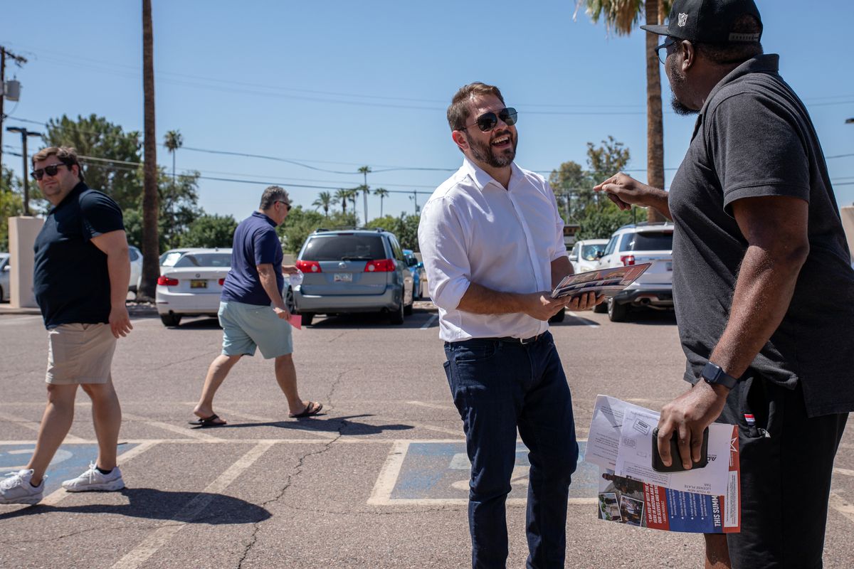 A candidate handing out election materials stands in a parking lot talking to a potential voter.
