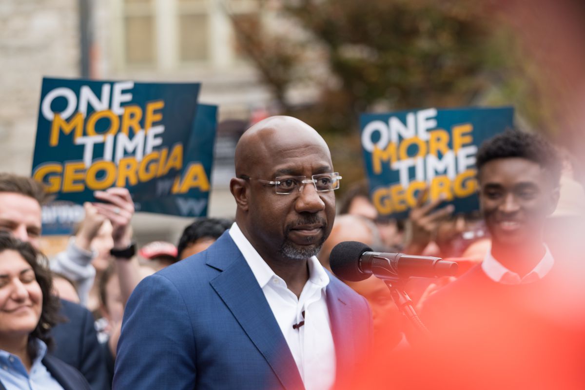Sen. Raphael Warnock speaks to a large crowd, some holding signs that read “One More Time Georgia.”