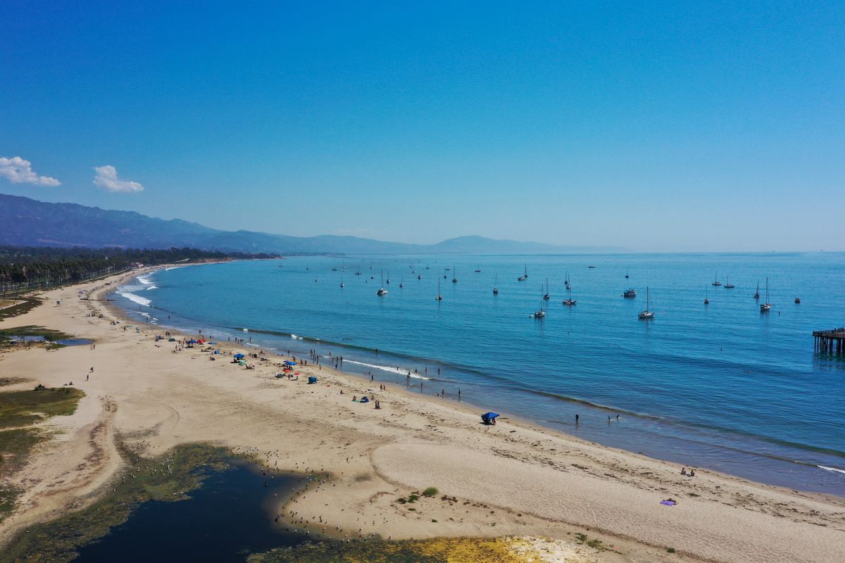 A long view shows a wide stretch of light sand California beach on a sunny day with small, faraway beachgoers along the edge, curving around a calm blue Pacific Ocean bay filled with small boats just offshore. In the background, mountains rise into an intensely blue sky.