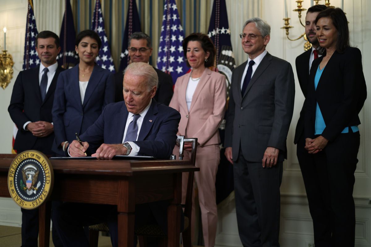 President Biden is seated as he signs the Executive Order on Promoting Competition in the American Economy. He is surrounded by a group of men and women looking on as he signs. 