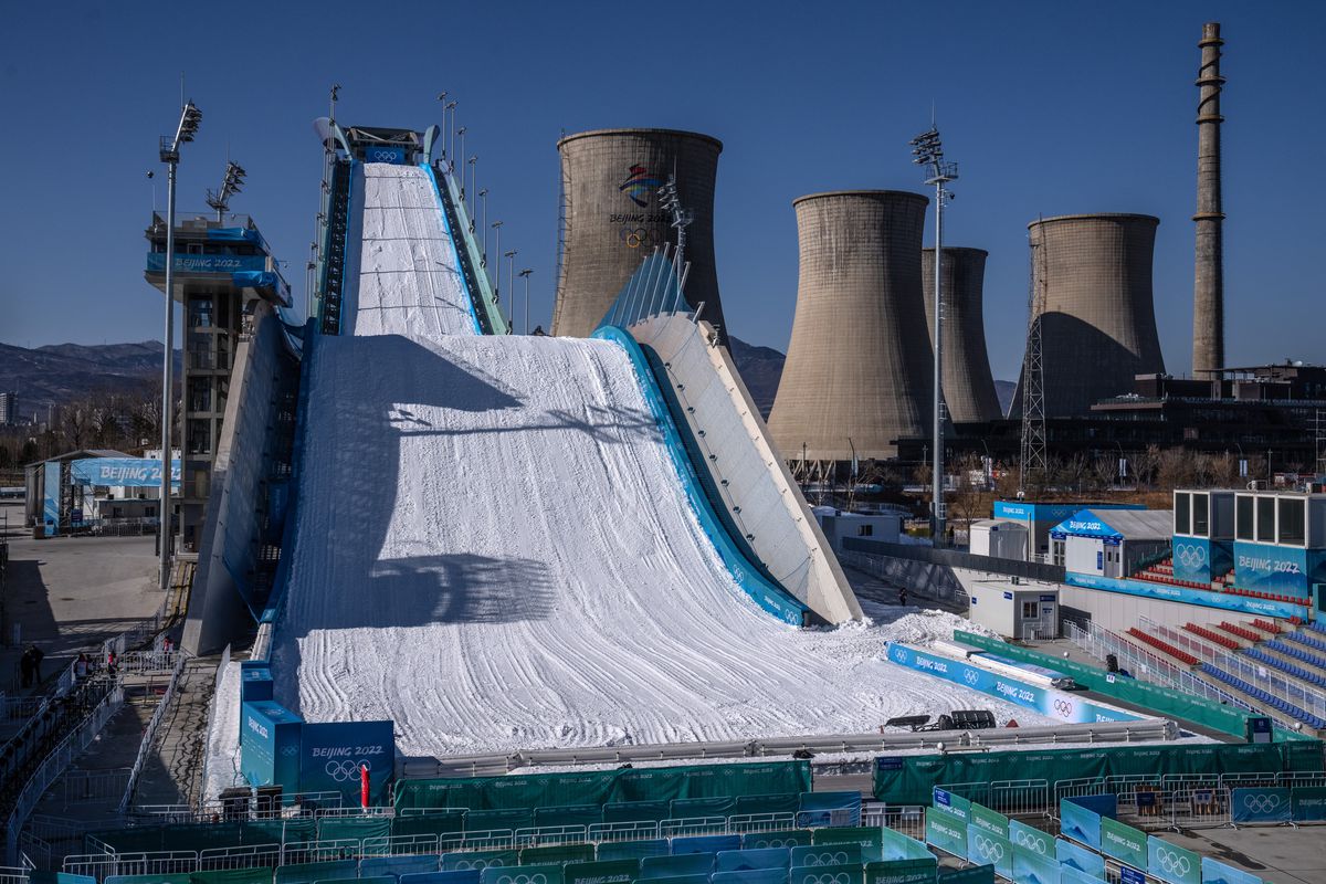 A long, curving ski jump covered in machine-made snow is pictured at daytime. Four large cooling towers loom just behind it.