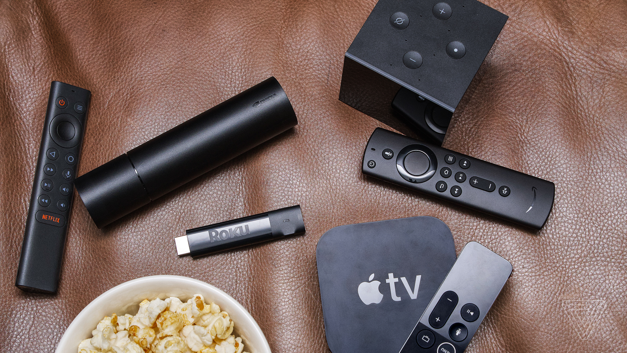 The Apple TV 4K, Amazon Fire Cube, Roku Streaming Stick, and Nvidia Shield streaming devices and remotes laid out on a brown leather couch next to a bowl of popcorn.