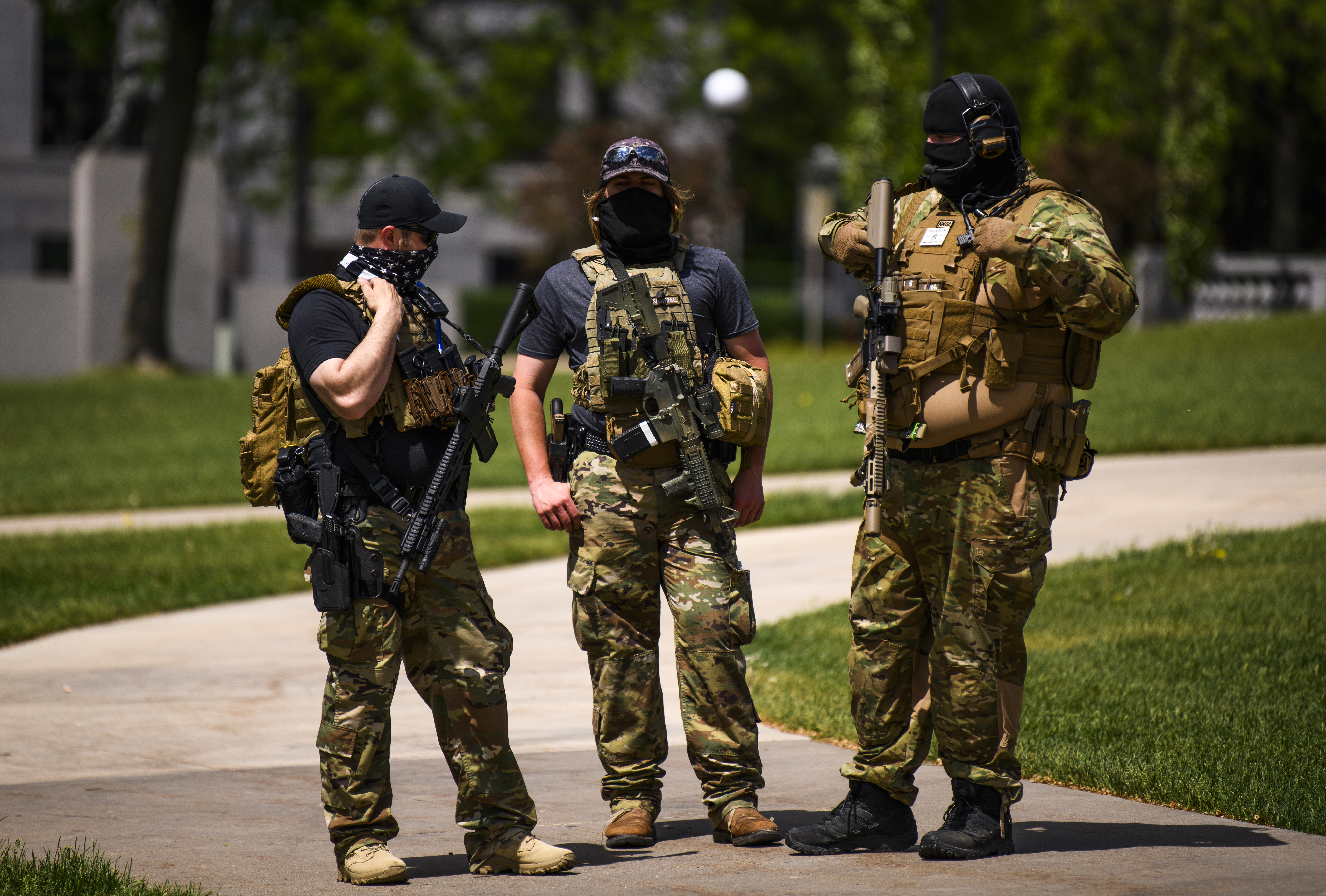 Three people in camouflage clothing stand with rifles and other gear in their hands and slung on their shoulders.