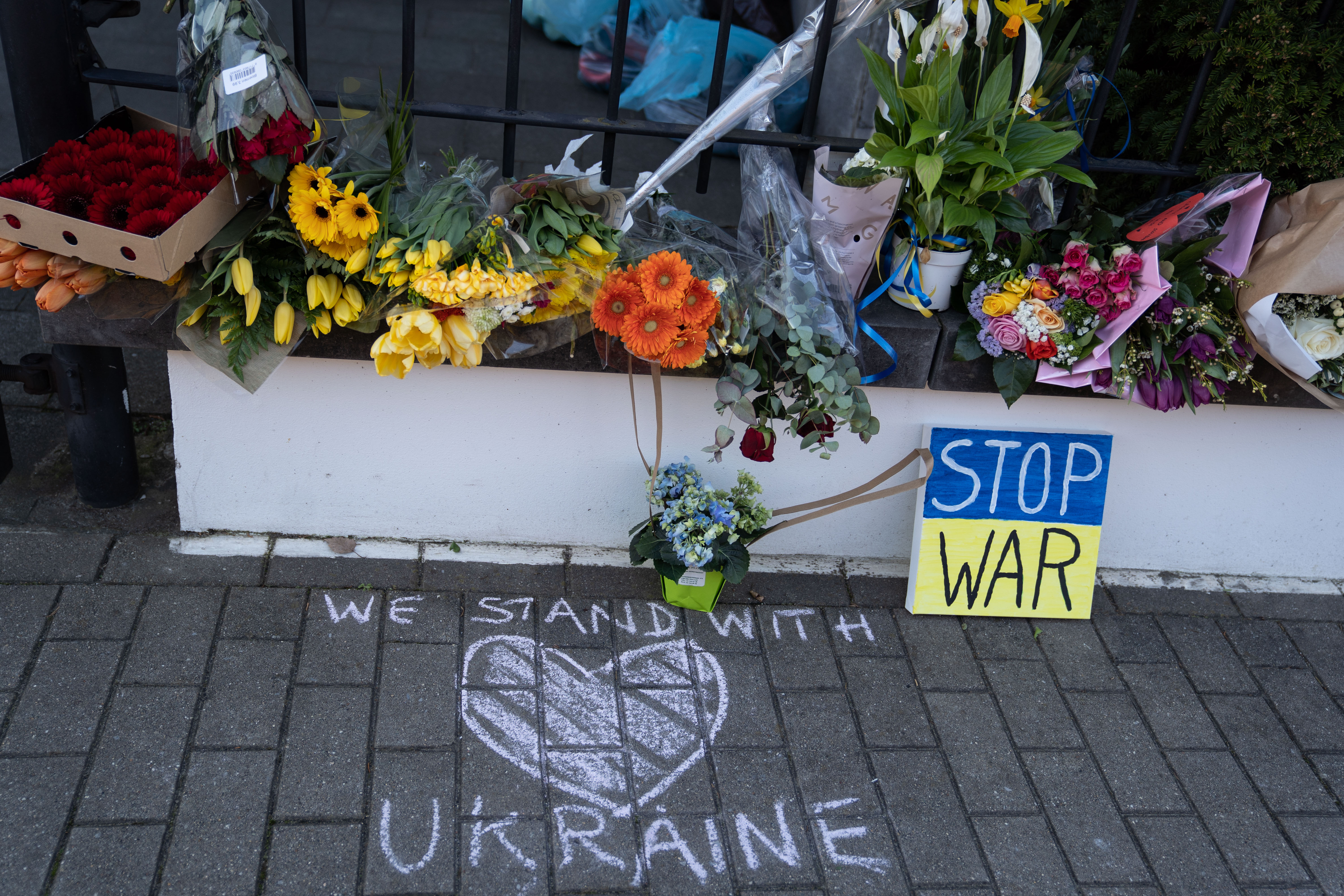 Bouquets of flowers outside the Ukraine embassy in Brussels accompany a small sign that reads, “Stop war,” and a chalked message that reads, “We stand with Ukraine” drawn with a heart.