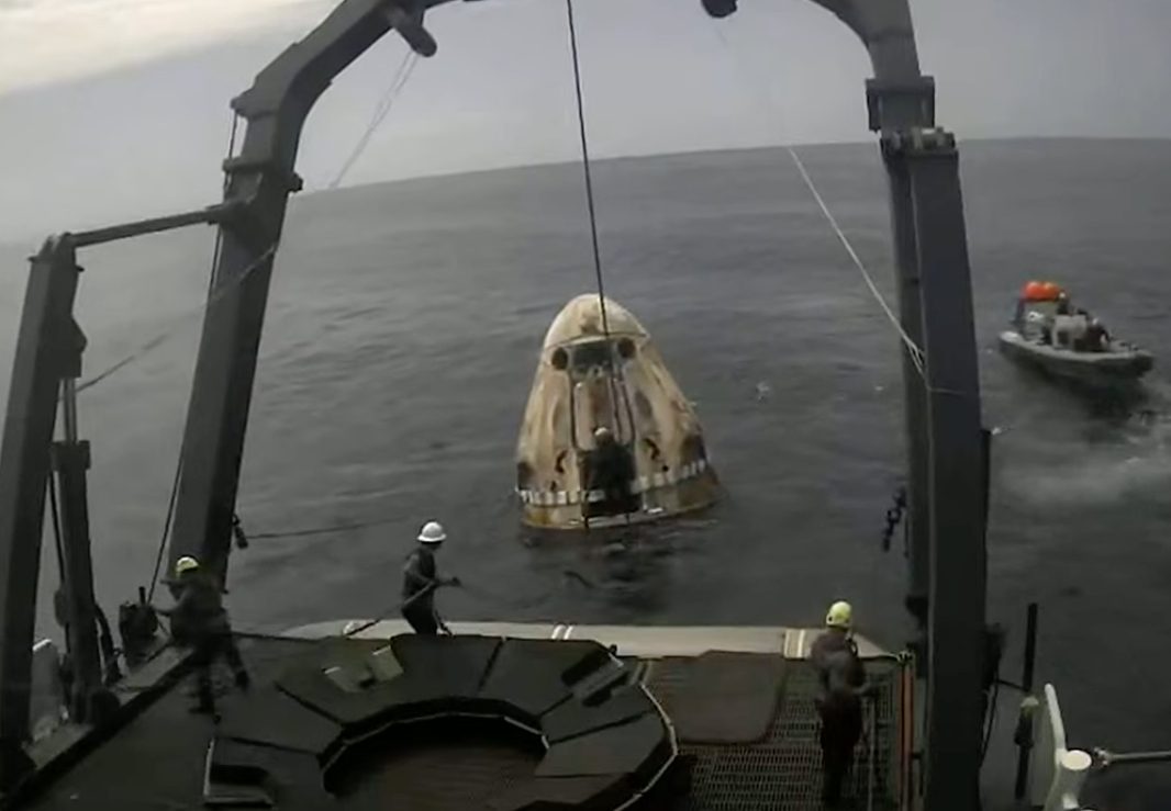 A crew dragon capsule bobs in the water, connected to a larger boat with tethers