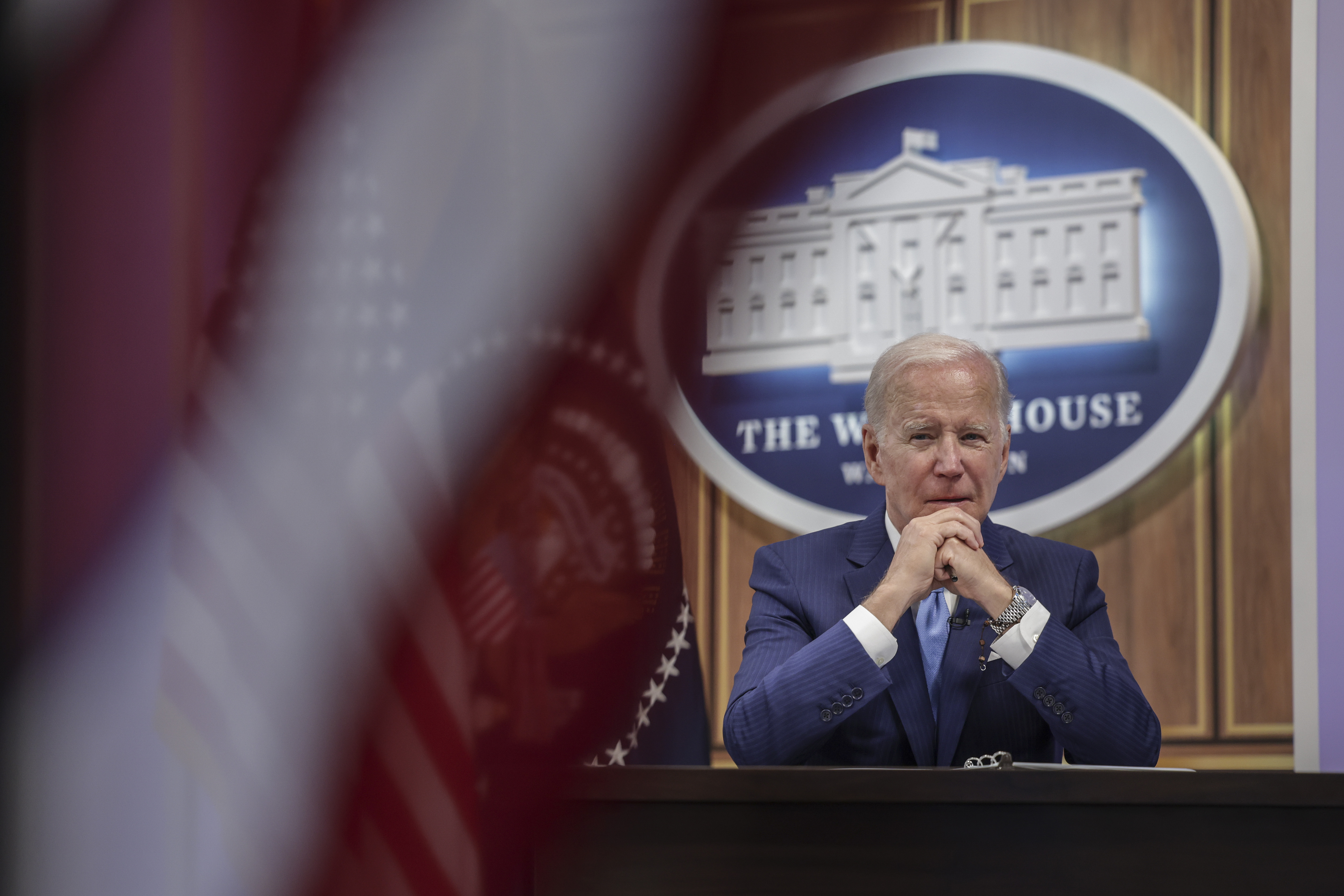 President Joe Biden listens to someone off-camera while sitting at a table with the White House logo behind him.