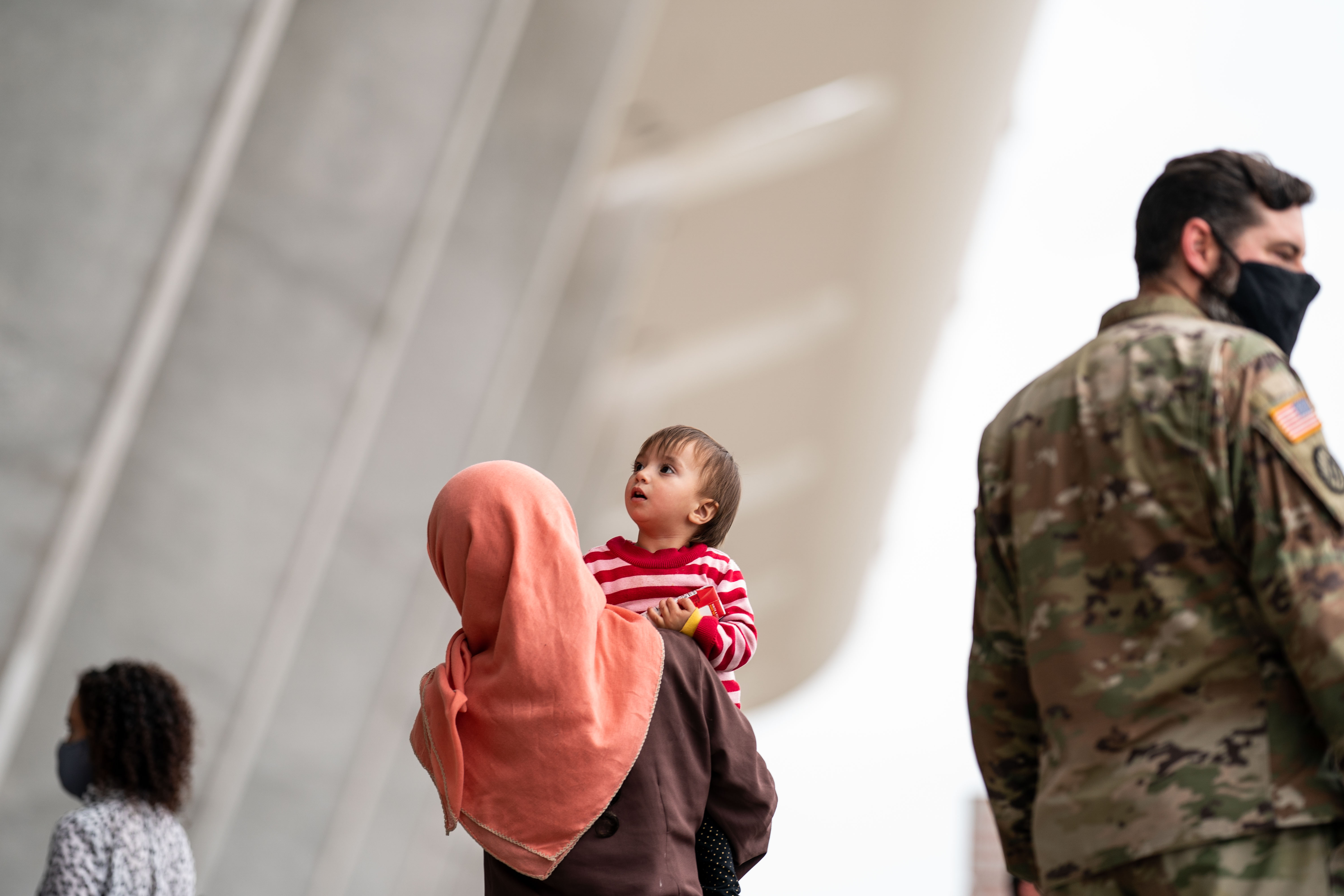 A person in a headscarf carries a small child past a soldier wearing camouflage.