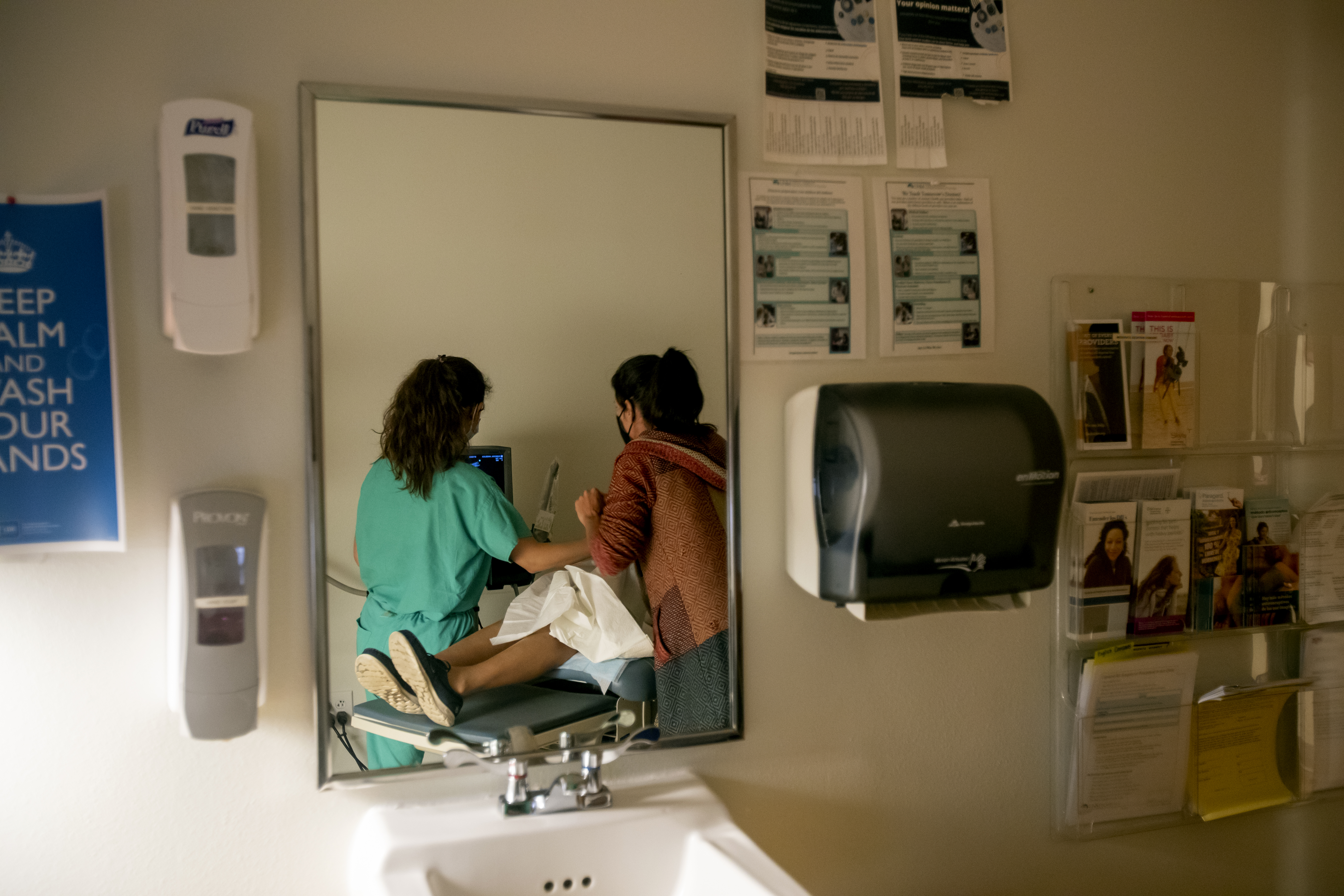 The reflection in the mirror above the sink in an examination room shows two women performing an ultrasound on a third.