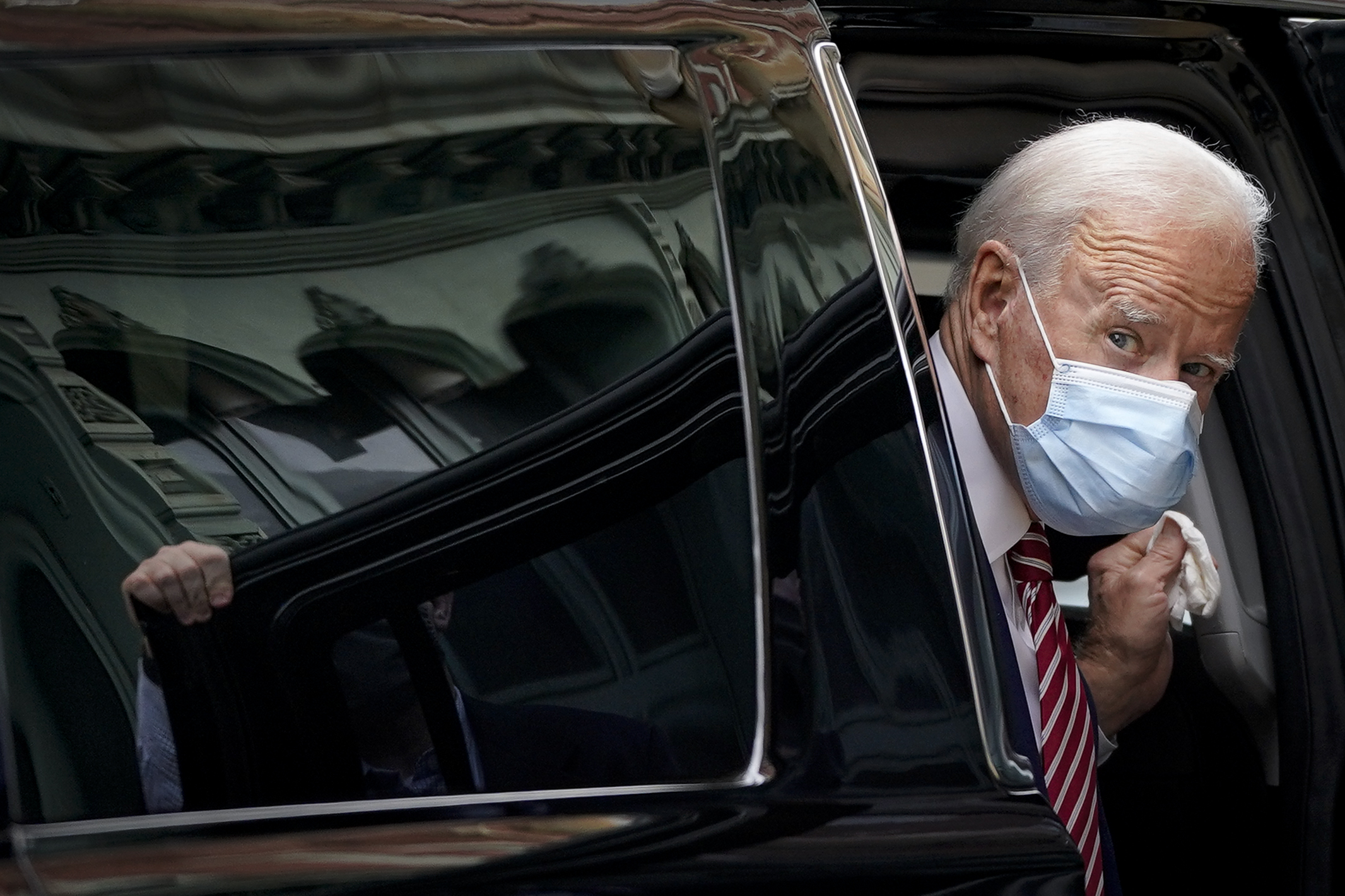 President Biden, wearing a blue mask and a dark suit, gets out of a black vehicle, looking past its door into the camera.