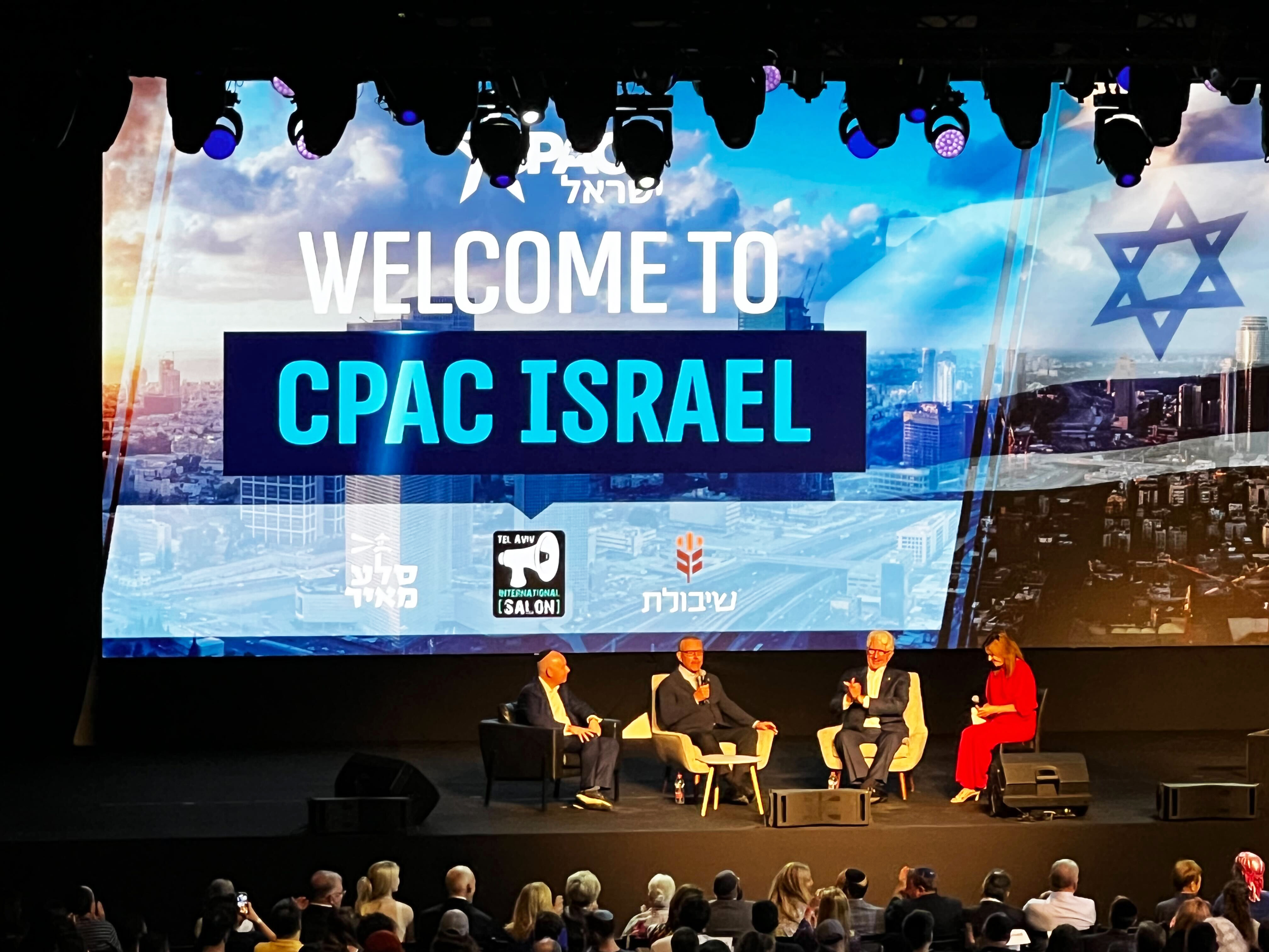 Four people sit on a stage. Behind them a large screen reads “Welcome to CPAC Israel.” 