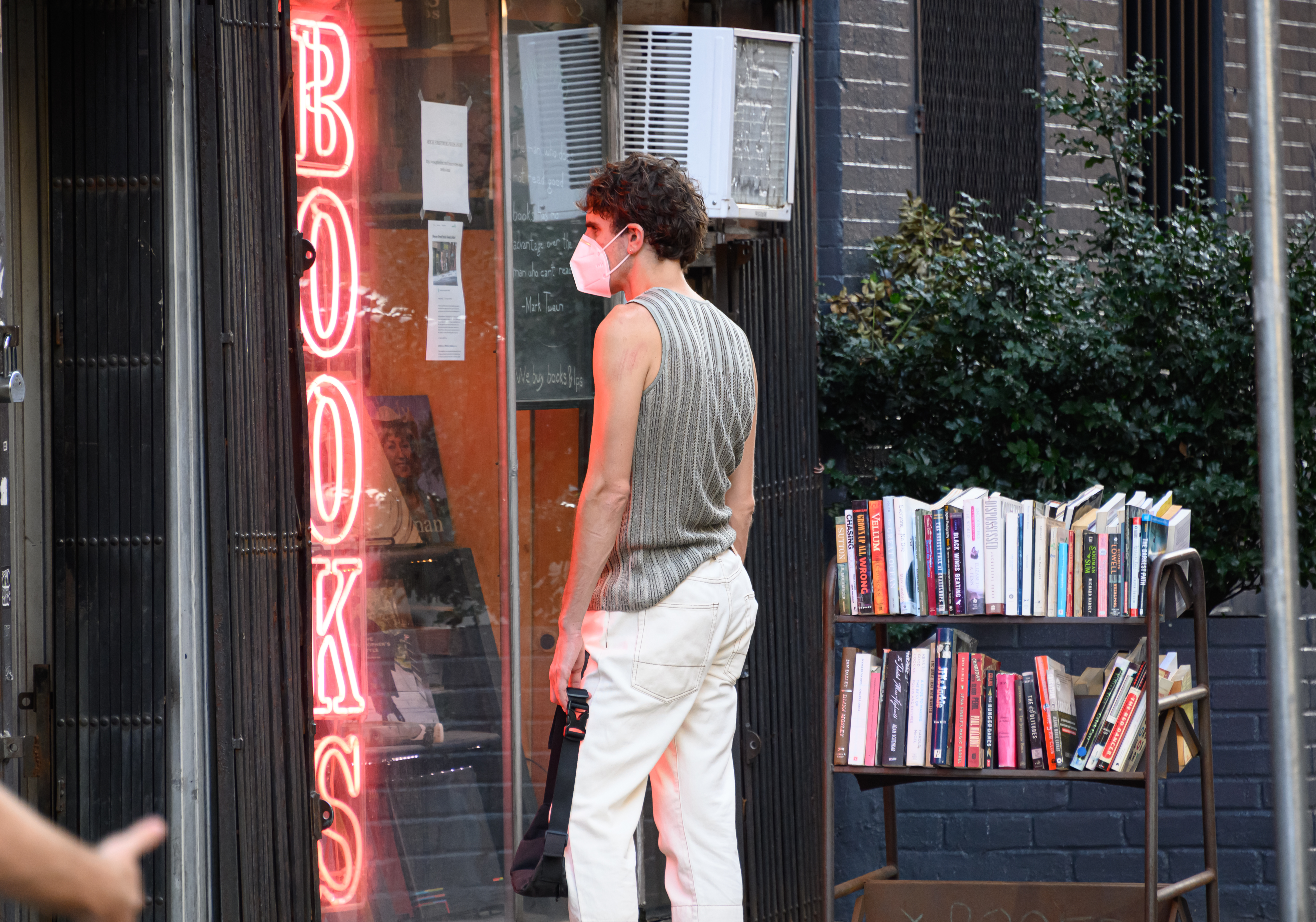A person standing beside a rolling cart of books while looking in a store window where there is a neon sign that reads, “books.”