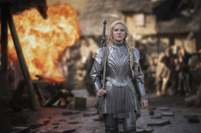 A women wearing armor and carrying a sword walks away from a burning building.