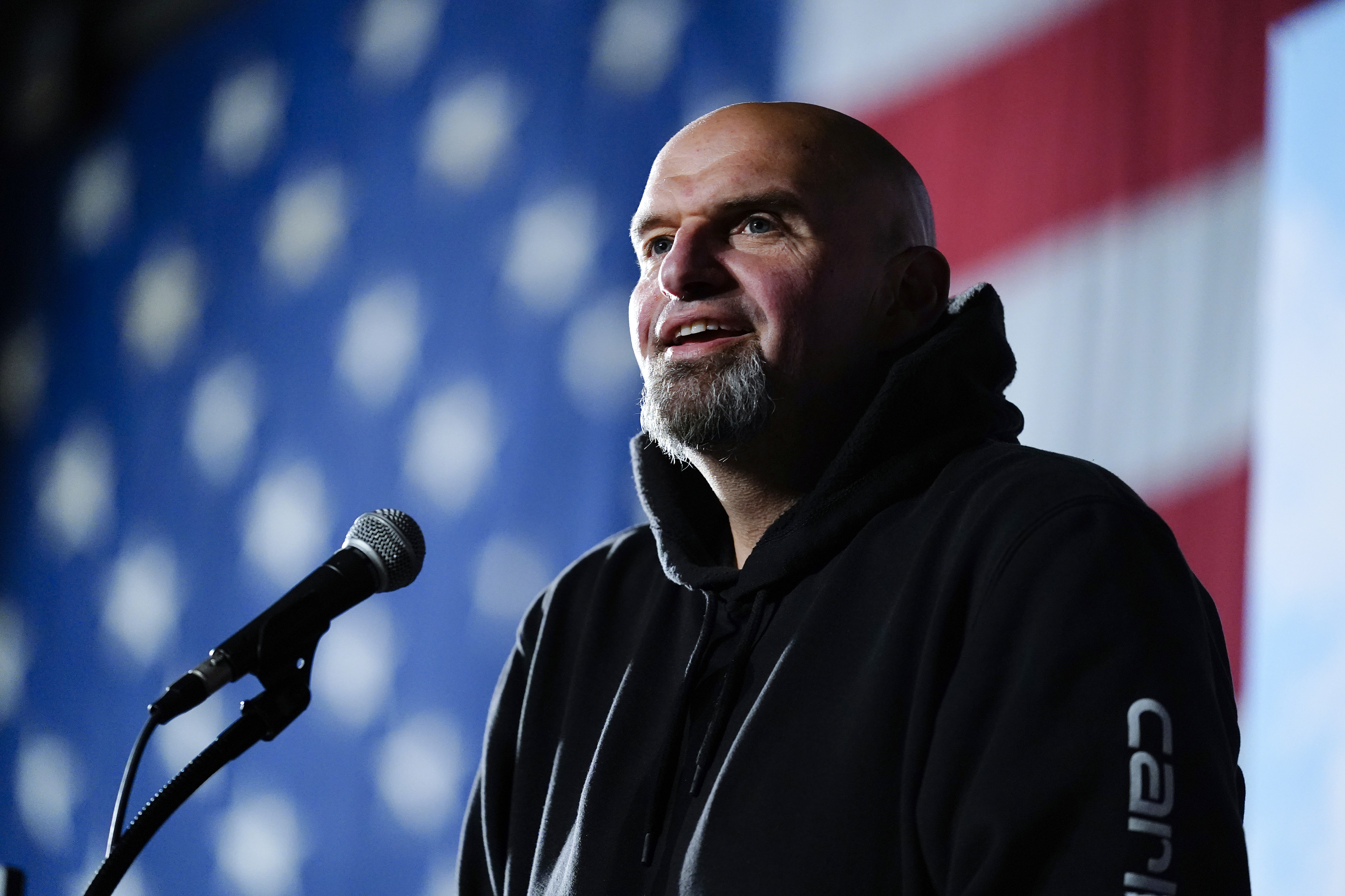 John Fetterman speaks into a microphone on a stage, wearing a black hoodie and smiling as he talks. Behind him is a large projection of the US flag.