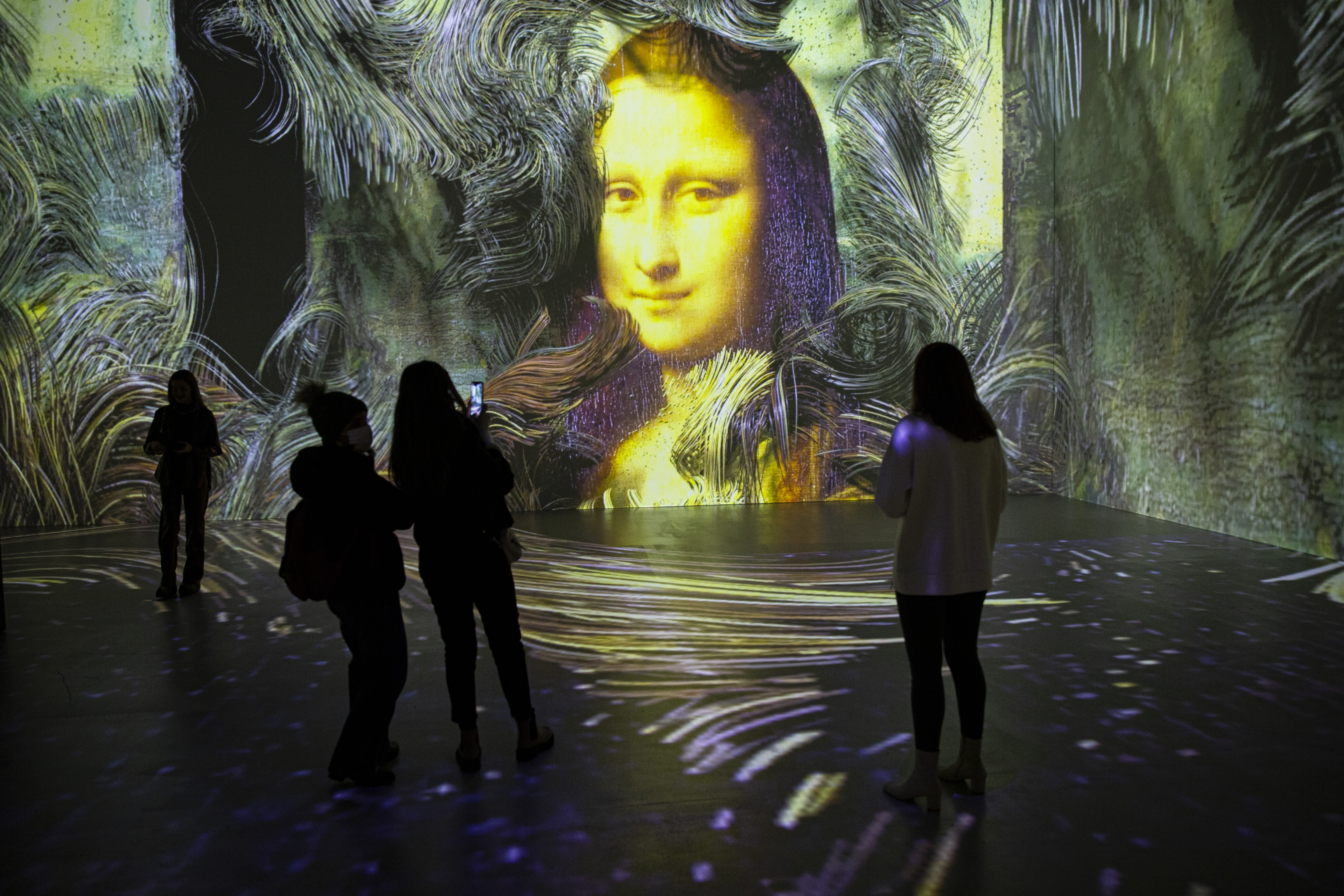 People stand in a dark gallery with a projection of the Mona Lisa surrounded by what look like swirls of paint on one wall.