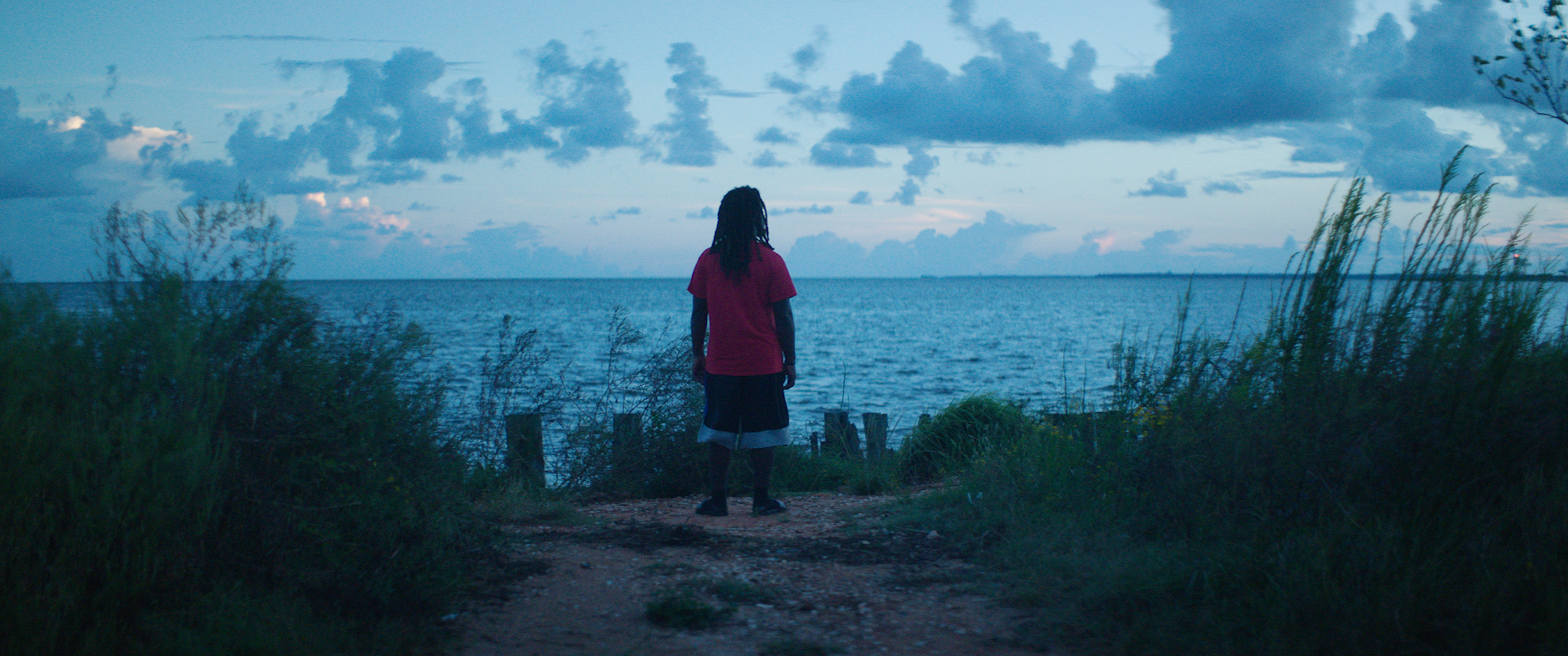 A Black man with dreadlocks stands facing a sunset over the ocean.