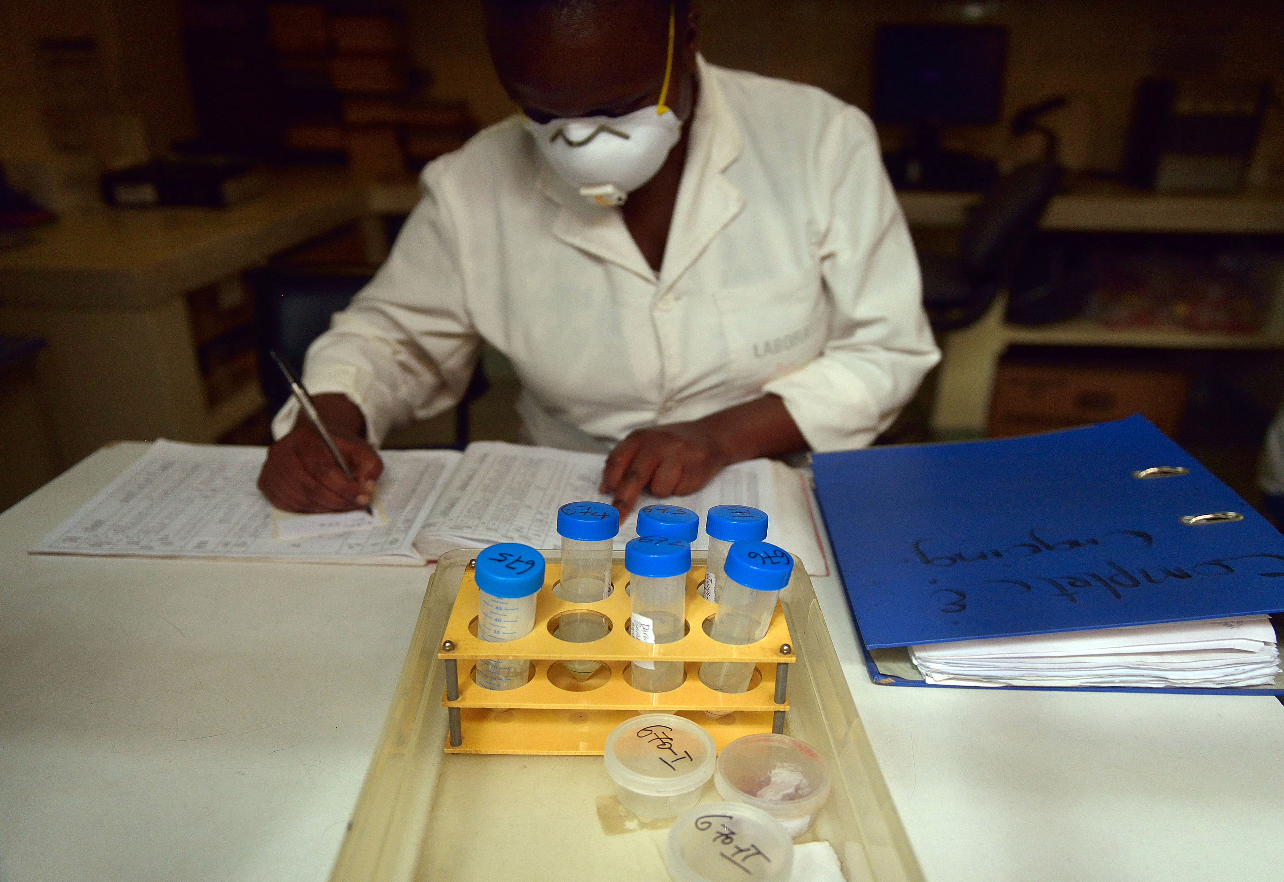 A person in a lab coat and mask sits at a table writing in a spreadsheet behind a tray holding vials of samples.