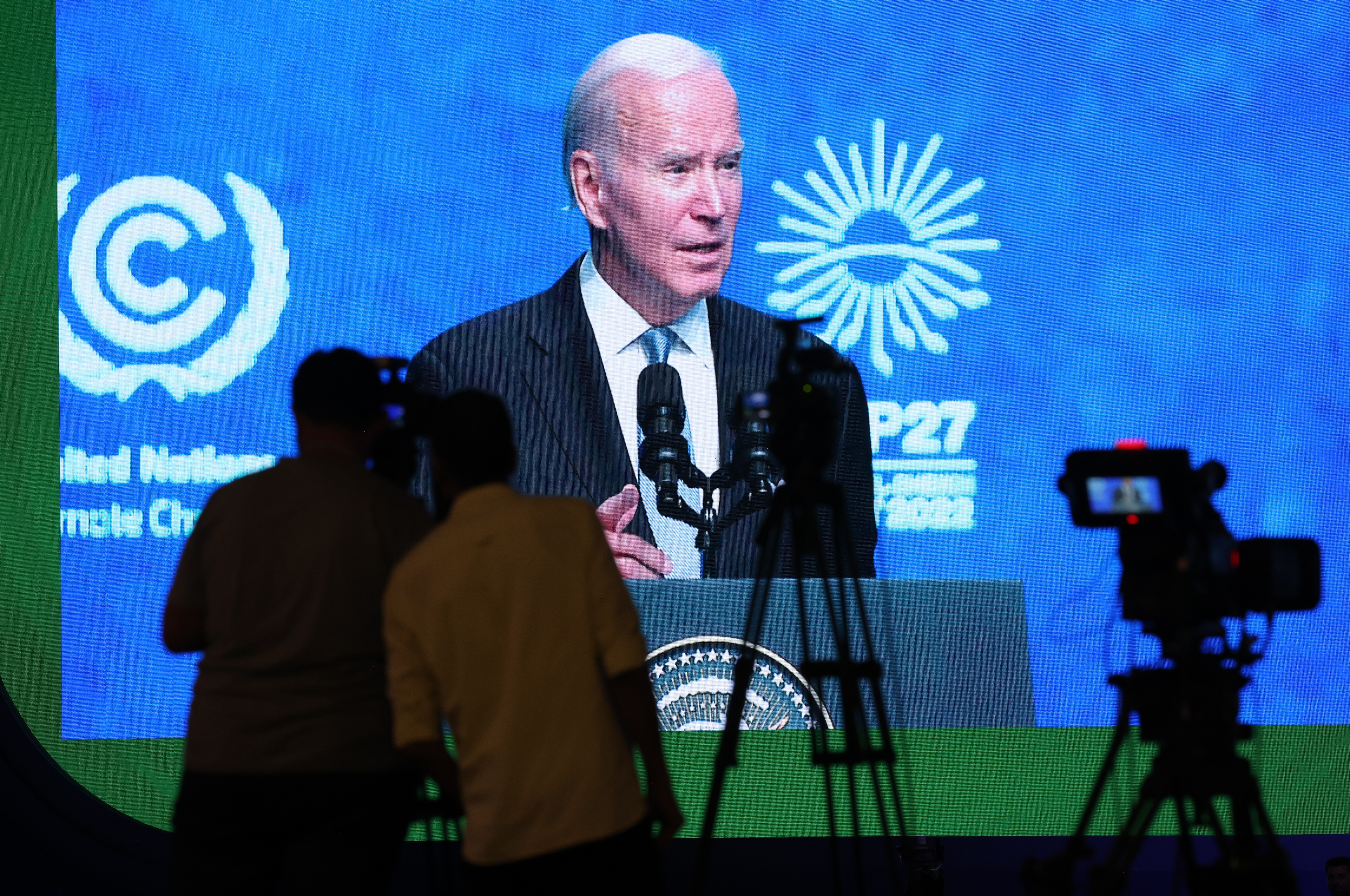 Two men watch President Joe Biden speak on a large screen. Behind Biden is a blue background with the UN climate change and COP logos. 
