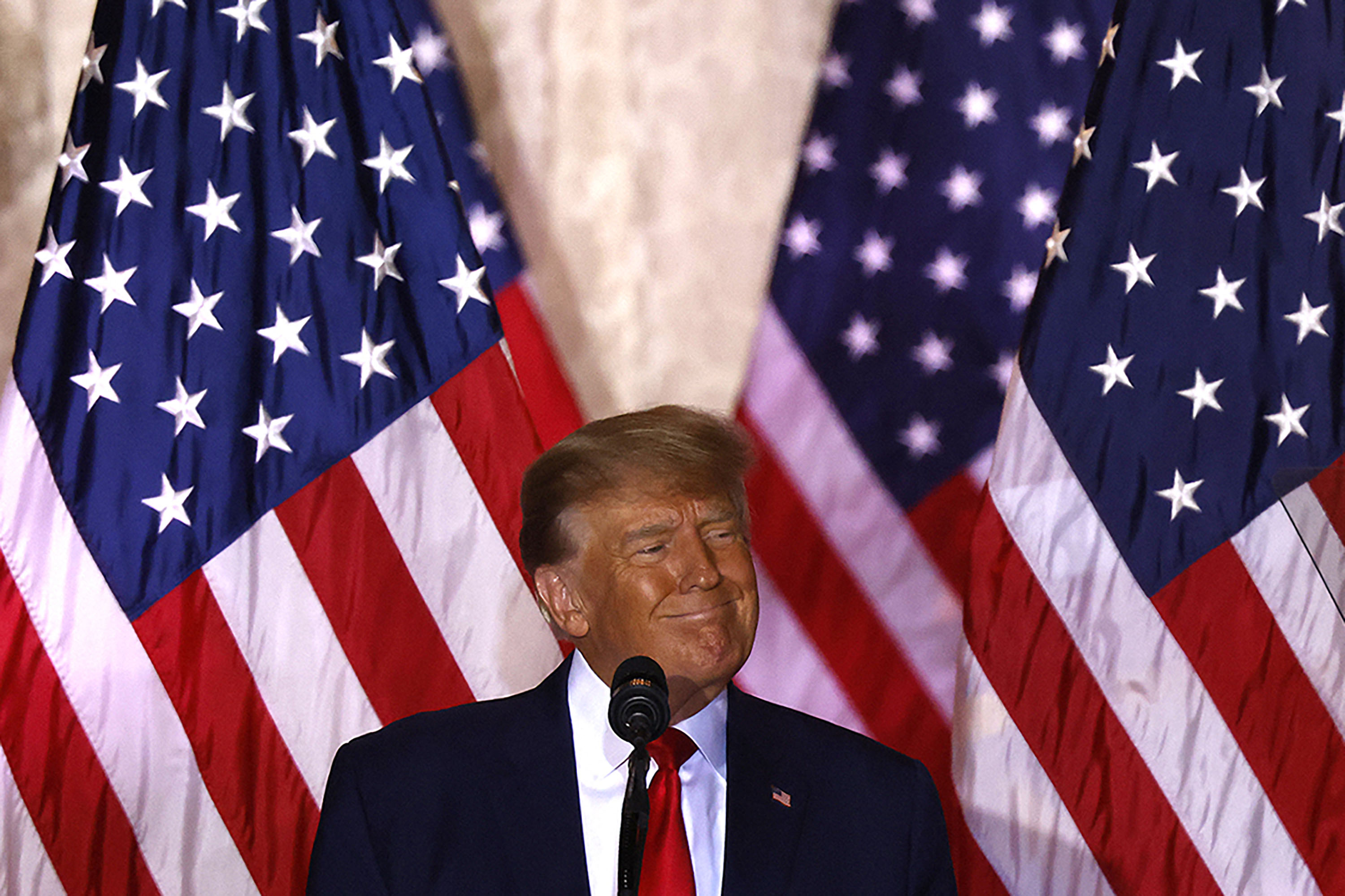 Donald Trump smirks at the camera, behind a microphone and in front of a wall full of American flags. 