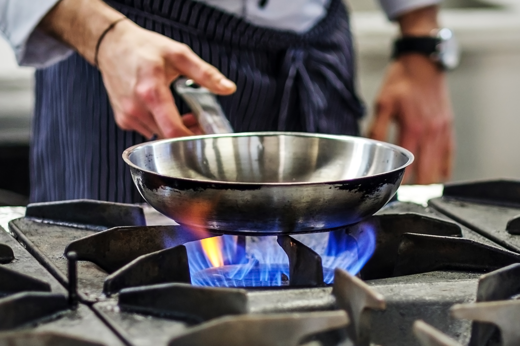 A chef holding a metal frying pan over a gas burner on an industrial stove.