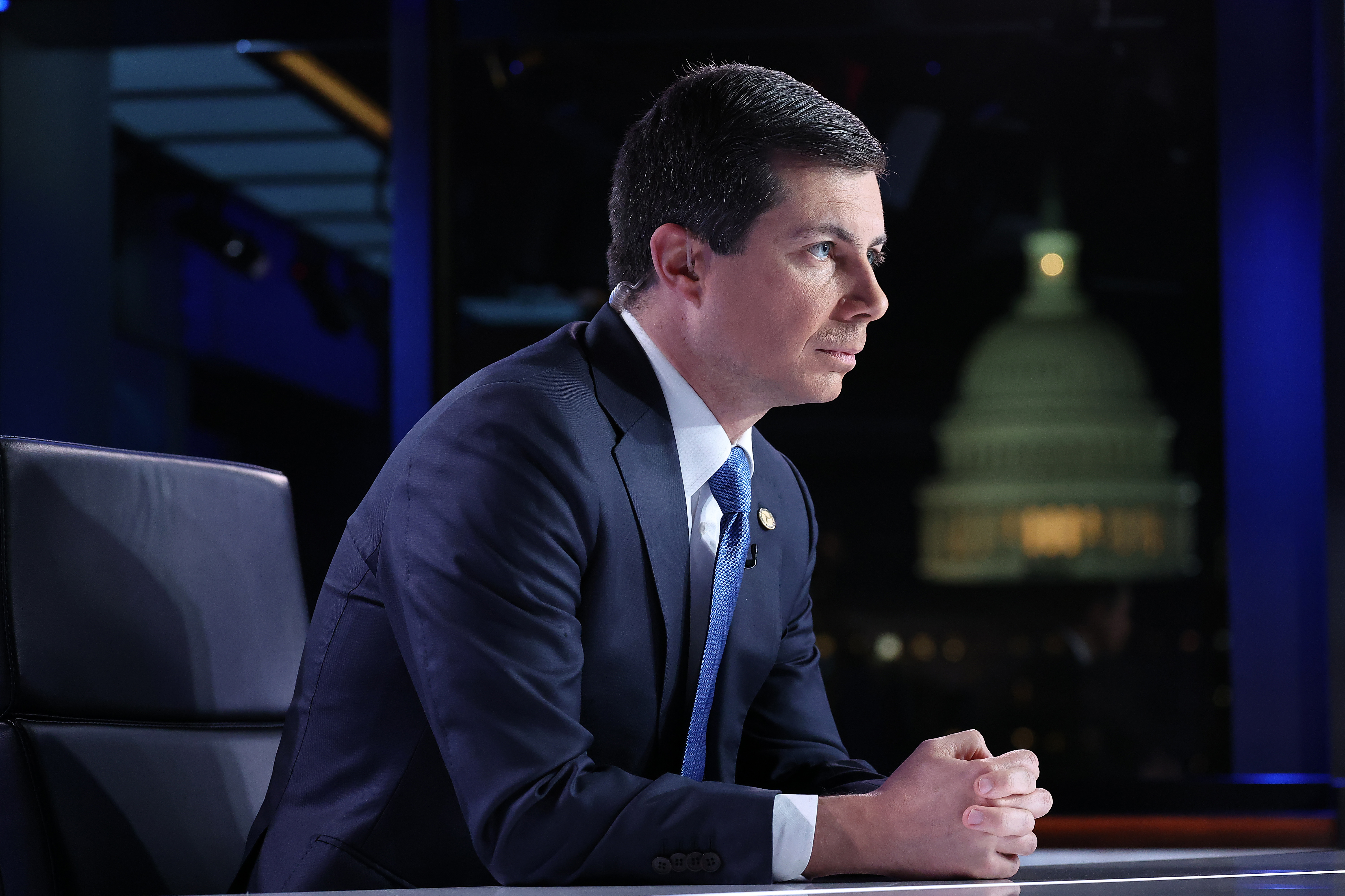 Secretary of Transportation Pete Buttigieg, in a blue suit and tie, listens with his hands clasped in front of him during a TV appearance.
