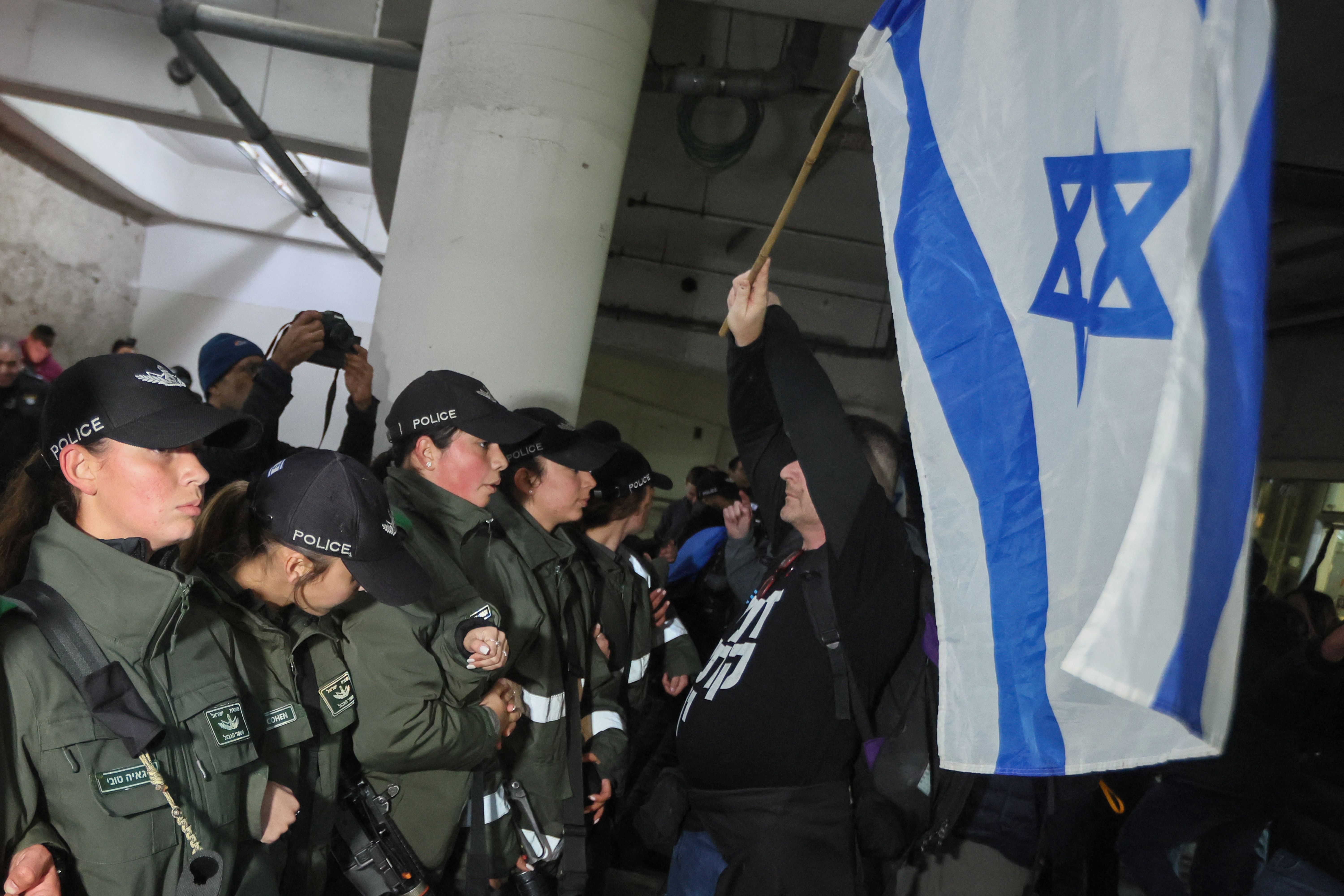 Security officers in green uniforms with rifles form a line pushing a group of protesters waving an Israeli flag back in a large indoor space. 