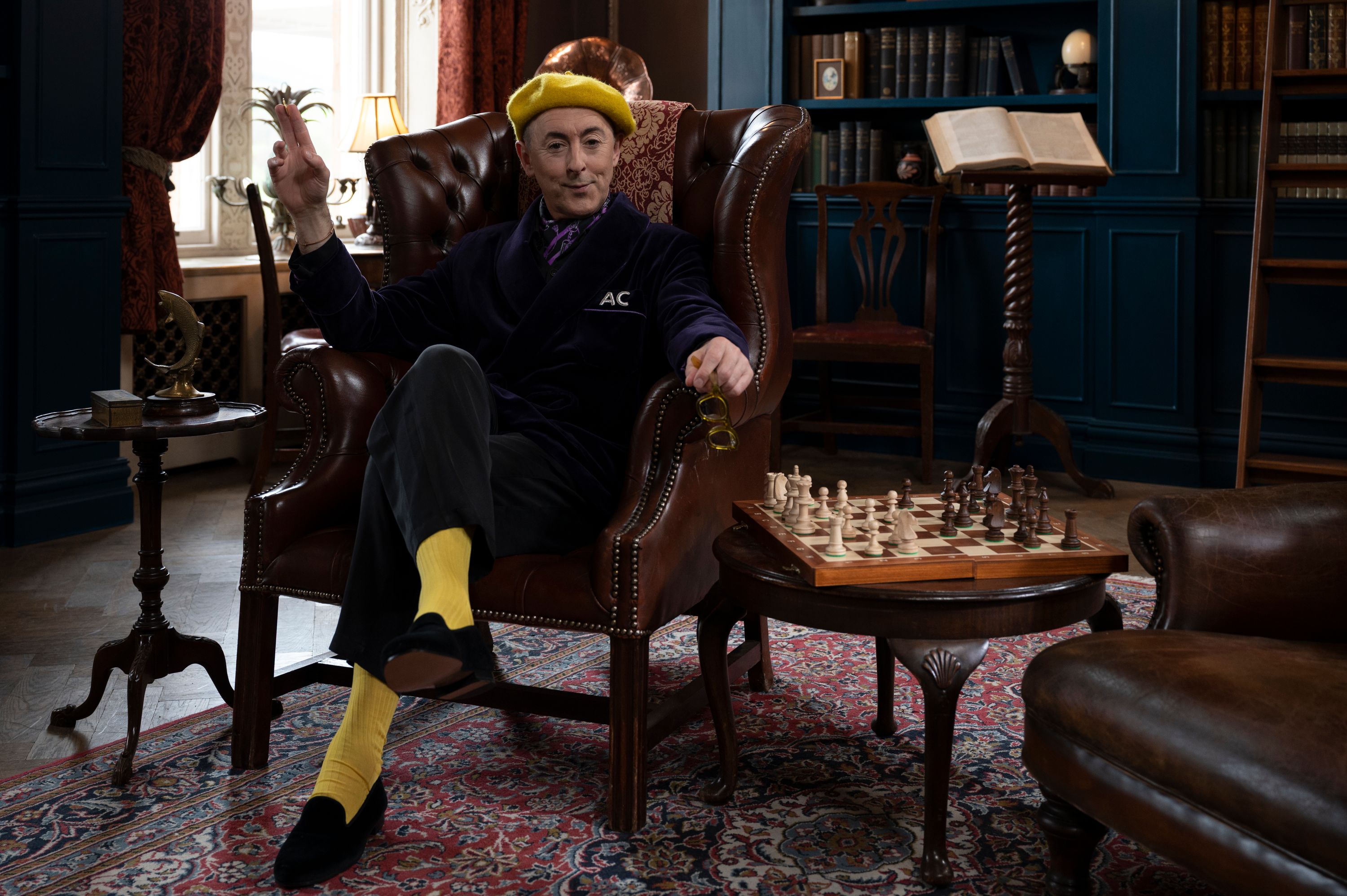 Actor Alan Cumming, in a dark suit with a yellow beret and socks, sits in an armchair with a chessboard on a nearby table. He hosts the TV show The Traitors. 