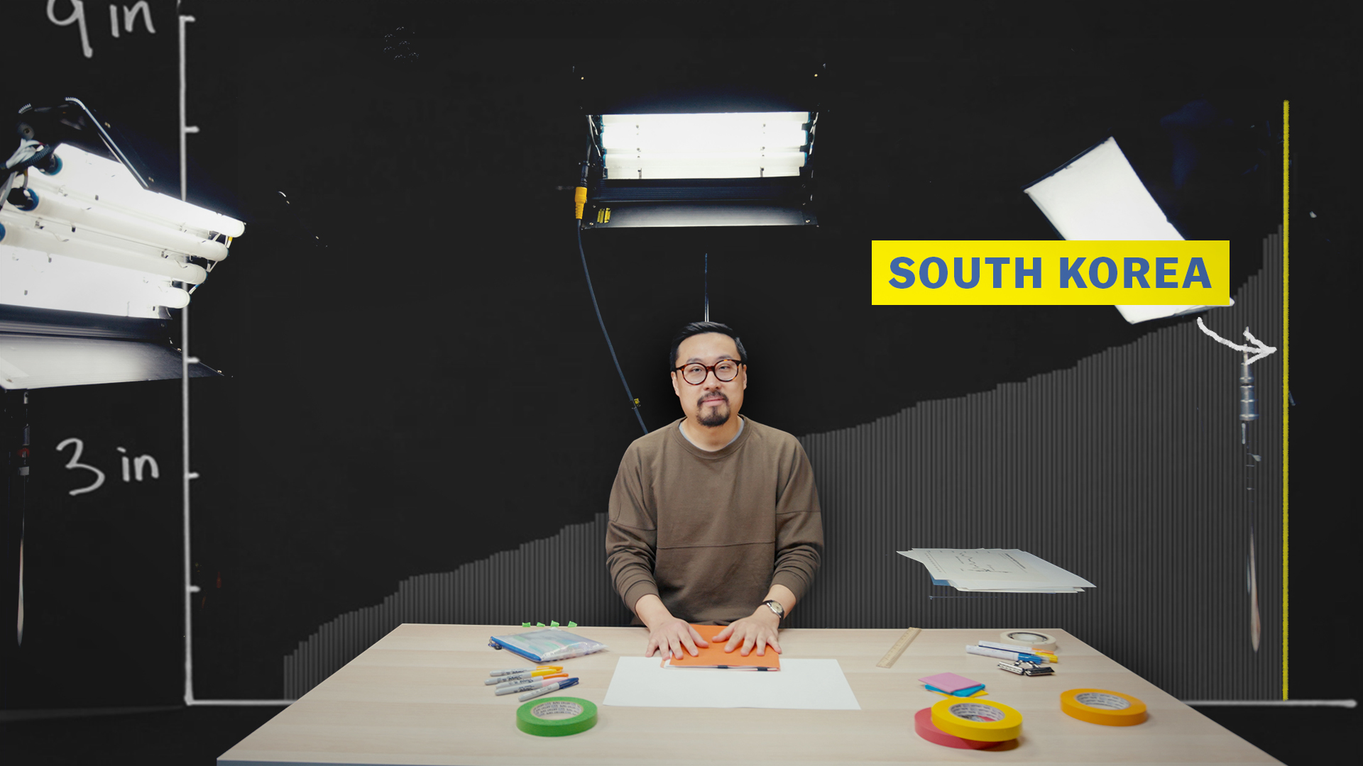 Vox journalist Alvin Chang sits at a table in a studio, surrounded by representations of the data he’s found on human heights over time.