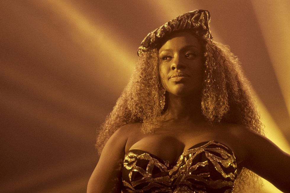 A Black woman onstage in a gilded bra top and matching hat. Light rays shine from behind her.