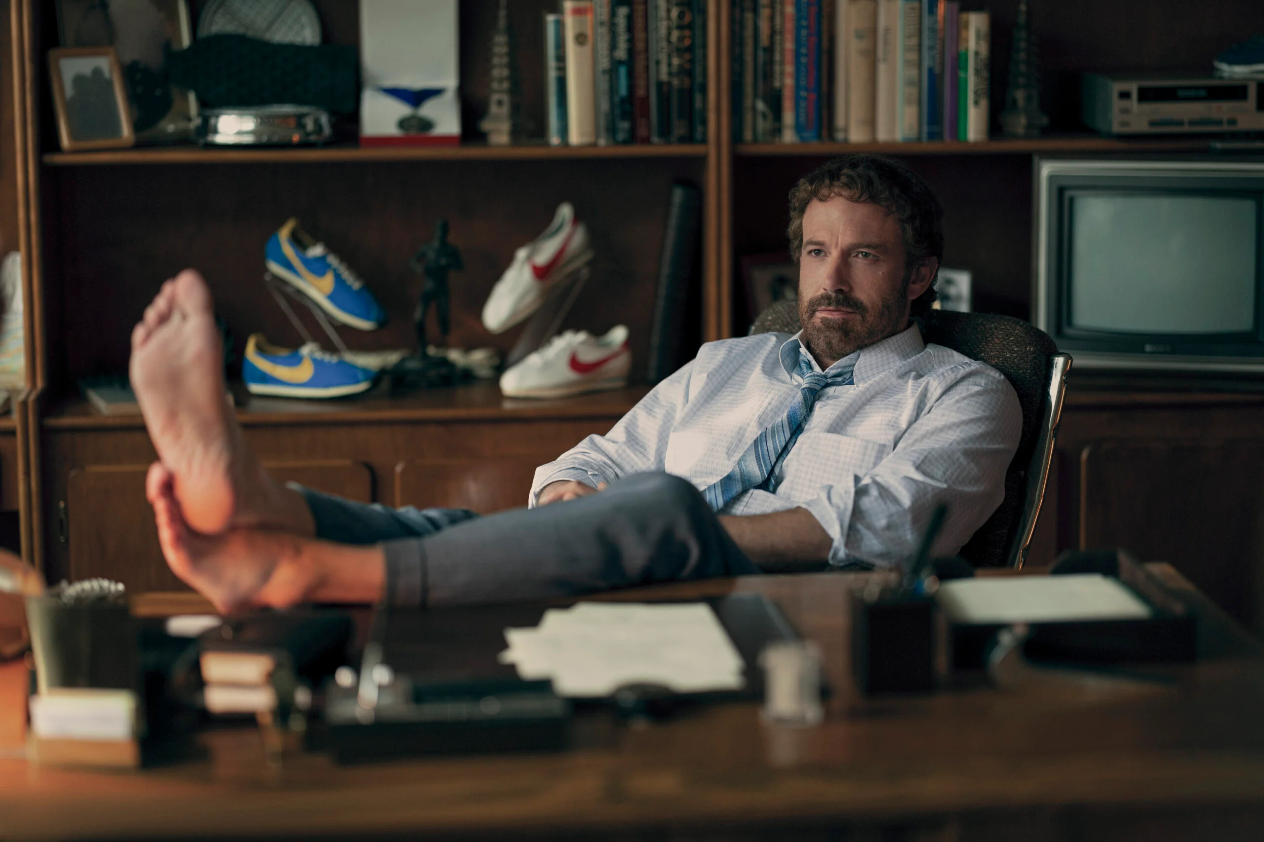 Ben Affleck, in a shirt and tie, sits with his bare feet propped up on a desk