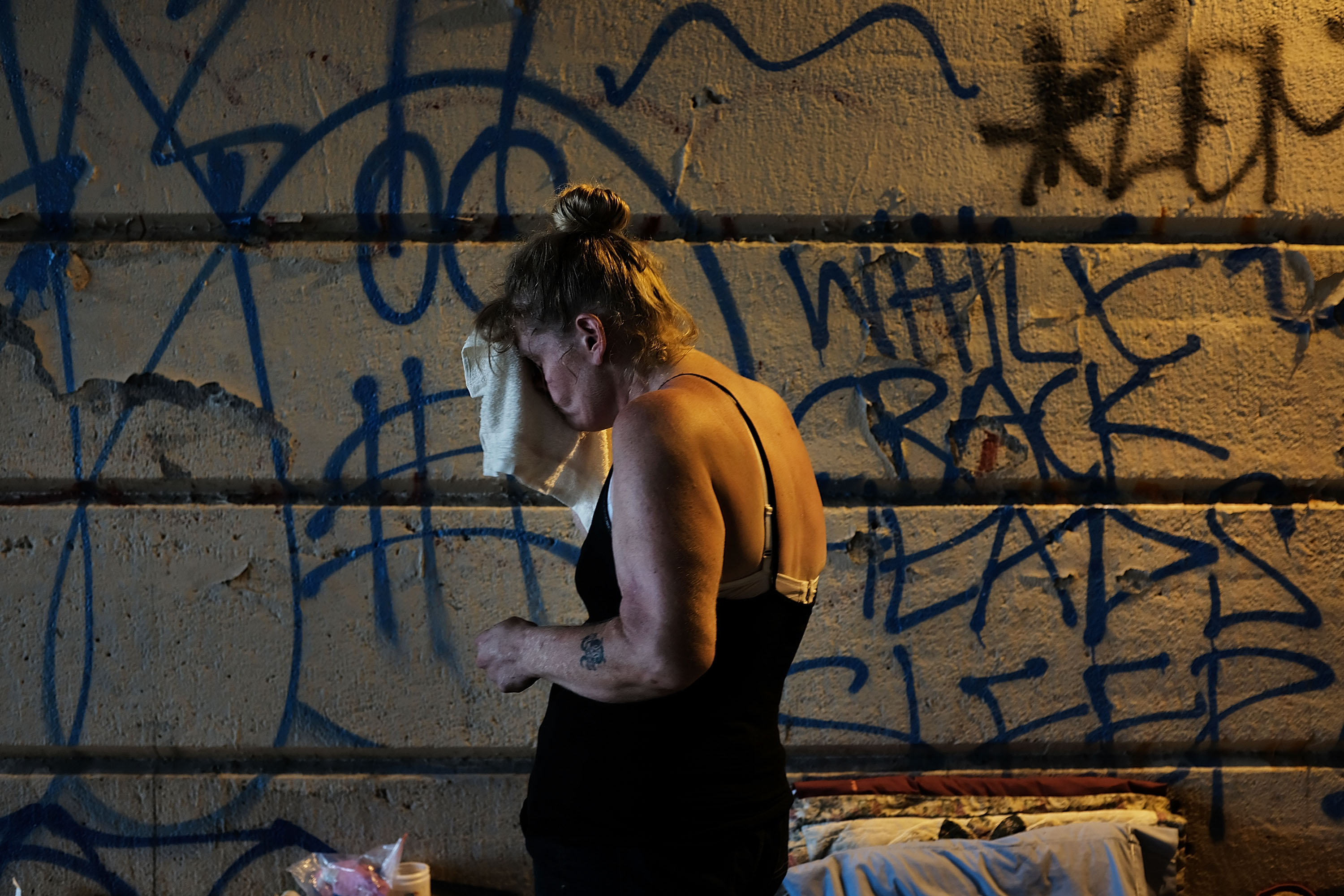 A woman stands in front of a graffitied wall holding a towel to her face.