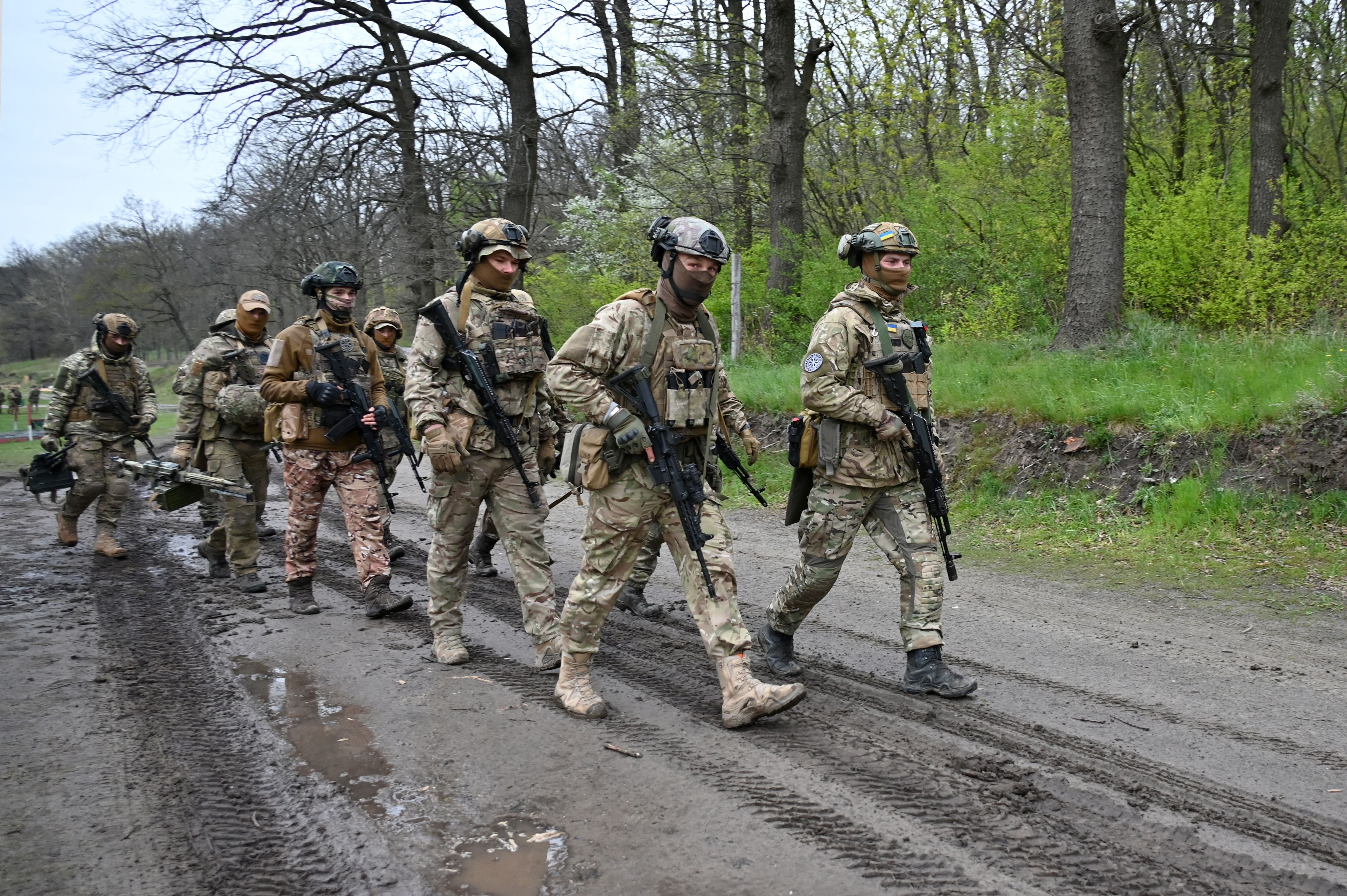 A group of soldiers in camouflage, helmets, and boots march down a muddy road, surrounded by trees.