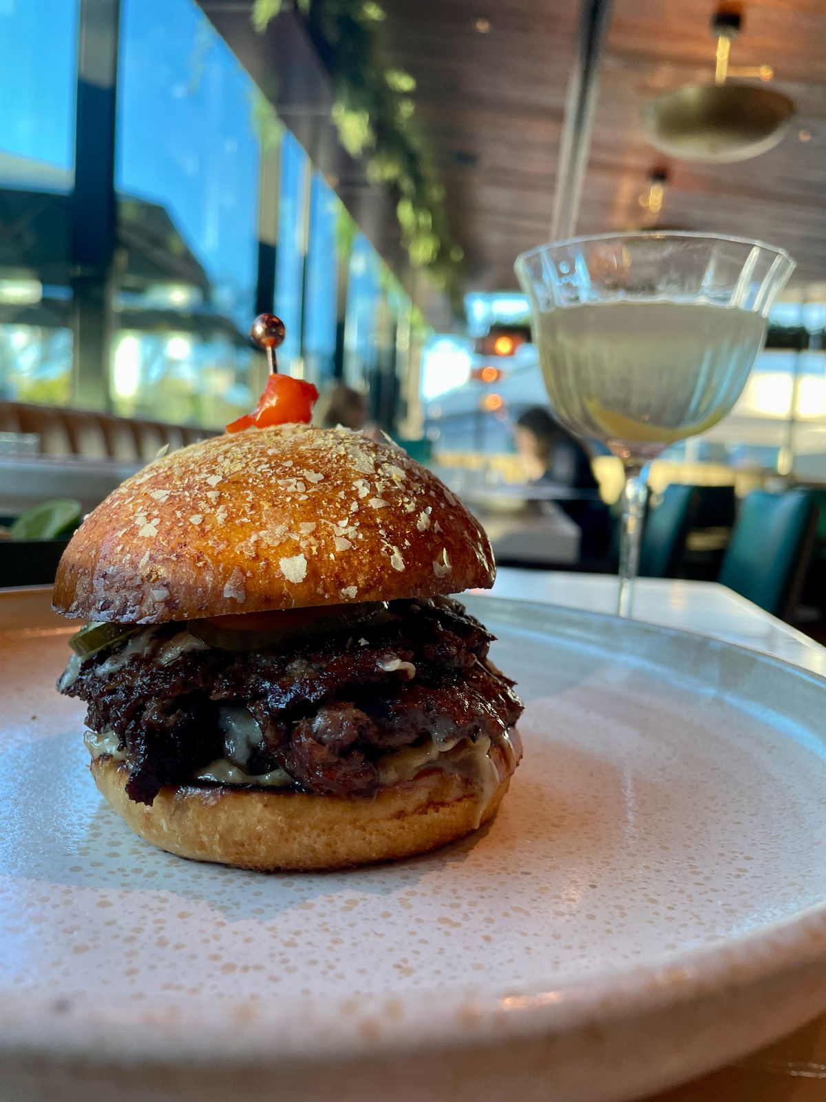 A double patty burger sits on a table with a martini glass to the right. Behind it are the brass and wood touches of a restaurant.