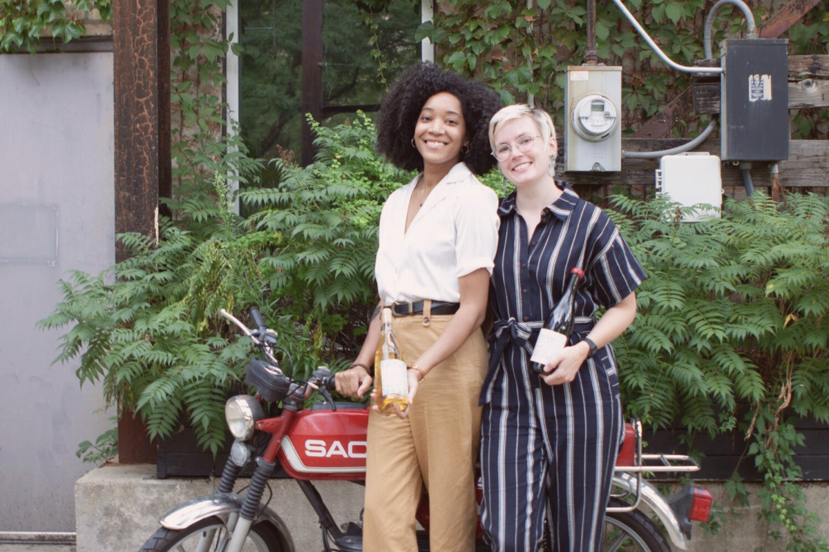 Two women stand together and pose, smiling, in front of a red vintage motorbike.