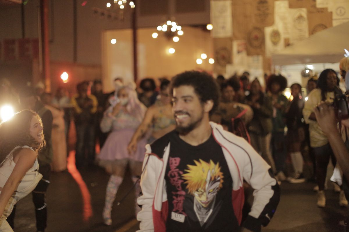 A young man wearing an anime t-shirt stands near a crowd of partiers and cosplayers. 