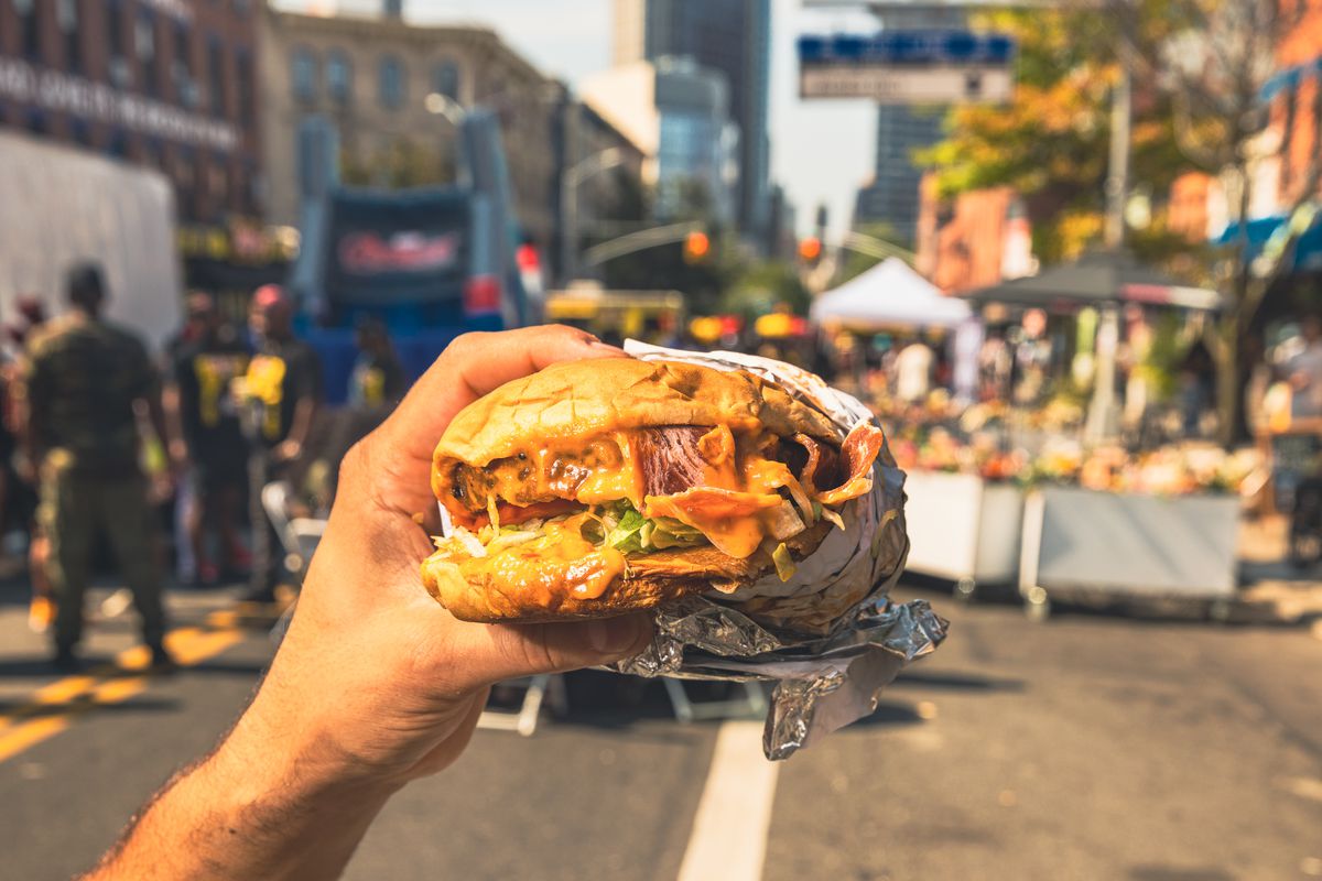 A hand clutches a vegan burger with meatless bacon and lettuce in front of a block party background.