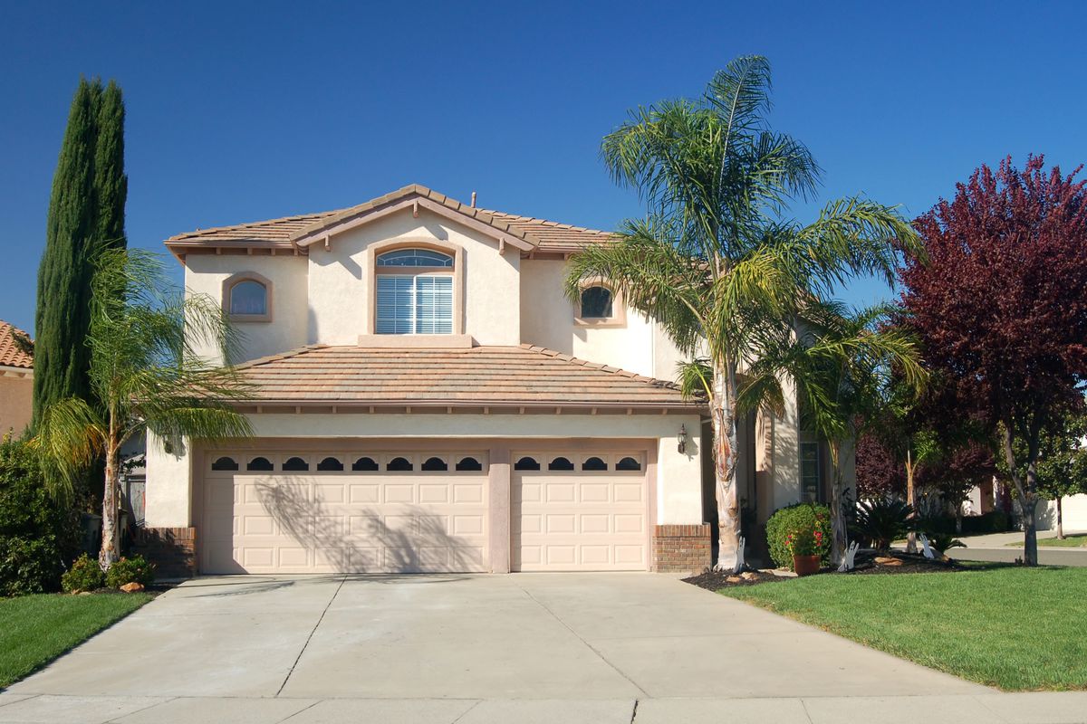 A Californian style two-story beige/light peach house, with a two-car garage, muted orange shingle roof, and a well manicured lawn with tall palm trees 