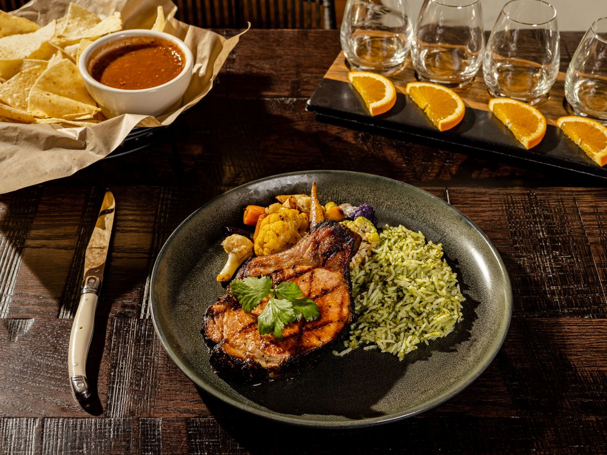 A dark wood table is set with a plate that holds a pork chop, cilantro rice, and mixed roast vegetables. In the background are chips and salsa, and a set of three glasses garnished with oranges.
