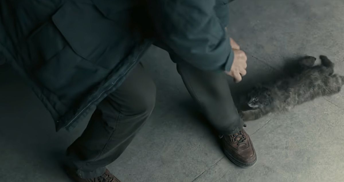 Otto (Tom Hanks) bends over to detach the claws of a pretty limp-looking CG cat that’s lying on its stomach on the ground of his garage, with its paws hooked into his pant cuff, in A Man Called Otto