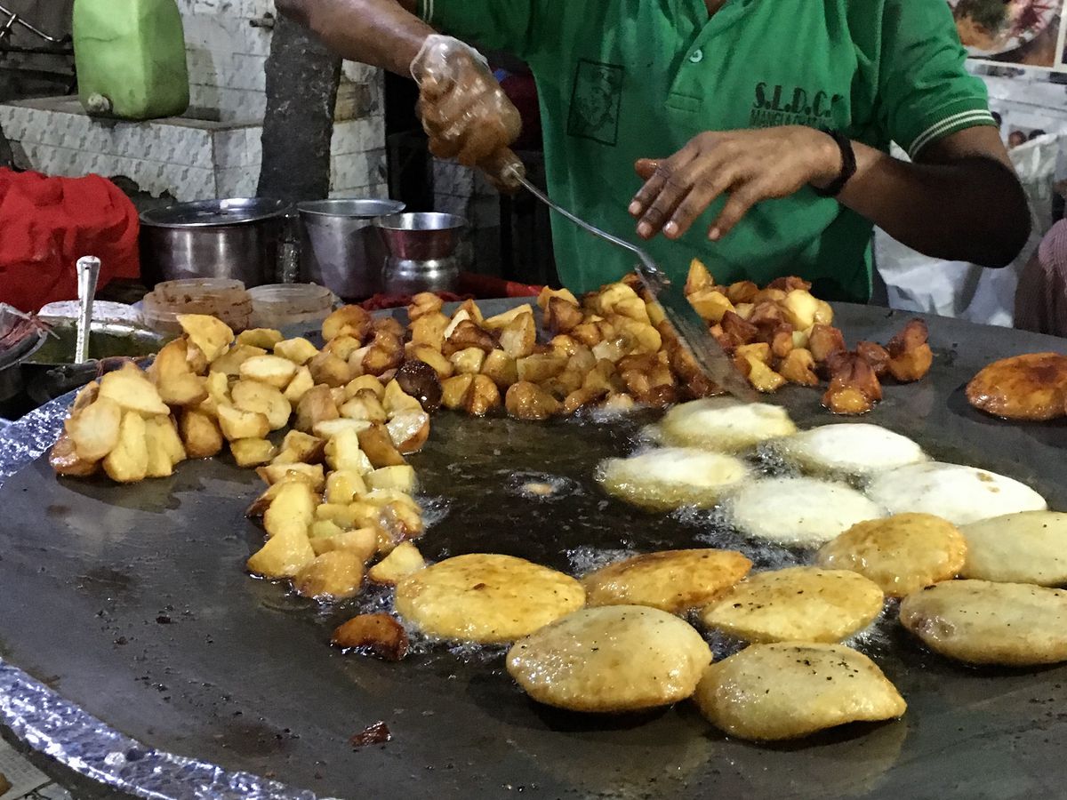A griddle with chopped potatoes and puffy kachoris sizzling, and a vendor stirring items behind the grill.