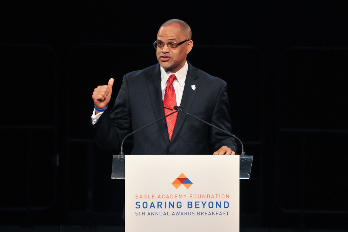 A man with glasses wearing a black suit and red tie gives a speech at a podium that reads, “Eagle Academy Foundation, Soaring Beyond, 5th Annual Awards Breakfast.”