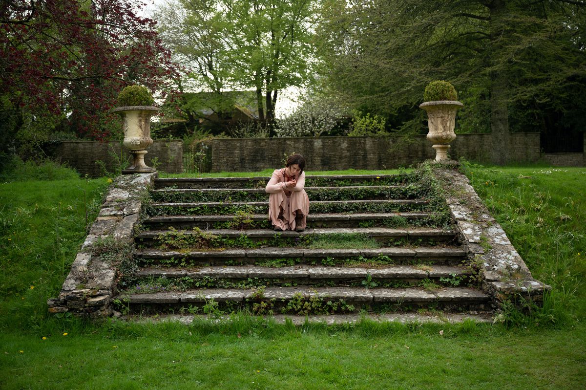 Jessie Buckley, in a long pink dress, sits on moss-covered stairs in a green landscape in Alex Garland’s Men.