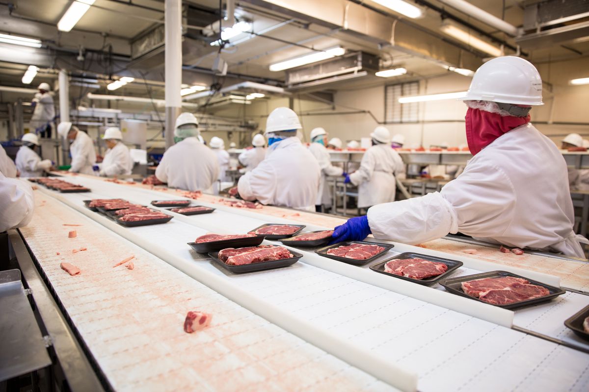 Meatpacking workers in full protective gear work an assembly line in a processing plant.