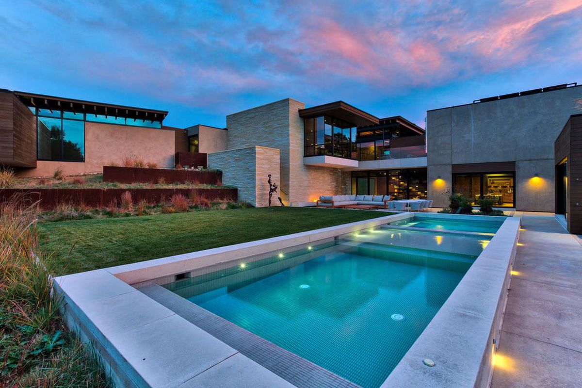 Exterior shot of of modern home from the backyard with swimming pool, lawn, tiered planters, and the cubic volumes of the stone, concrete, and glass home in the background.