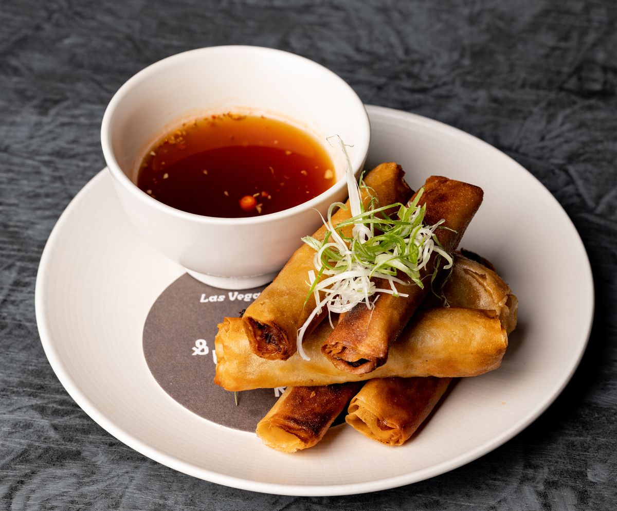 Stack of fried spring rolls topped with scallions with a bowl of rusty brown dipping sauce next to it.