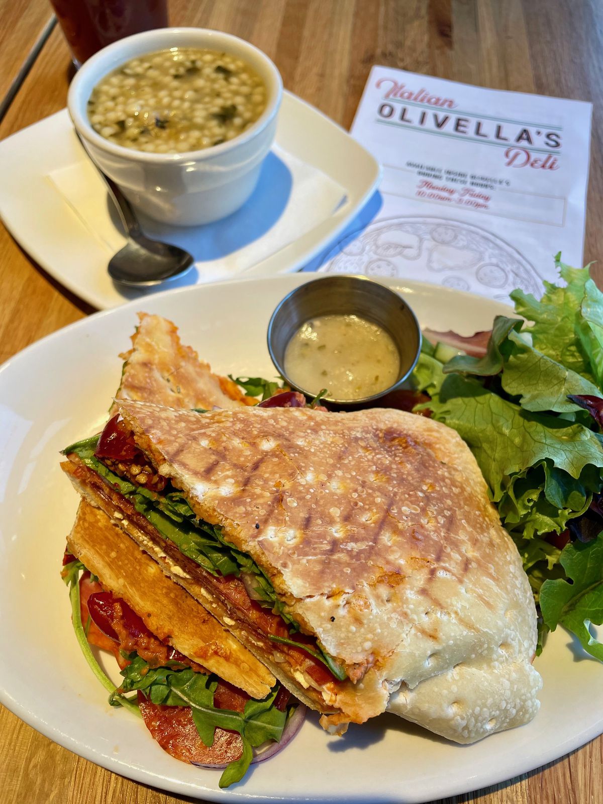 In the foreground, on a white plate, is a panini with a side salad. In the top left, a cup of Italian wedding soup with a spoon. In the top right, a menu that reads “Olivella’s Italian Deli.”