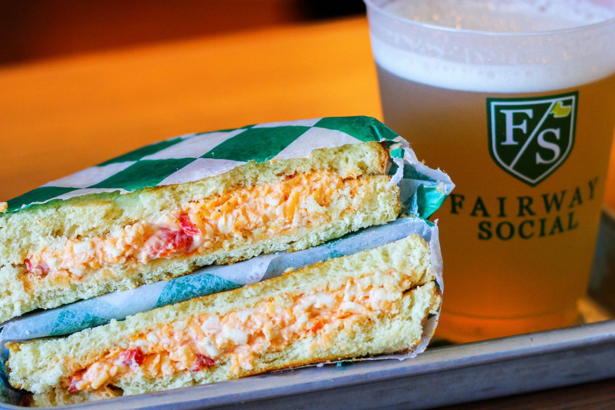 A pimento cheese sandwich cut in half sits beside a plastic beer cup filled with beer at Fairway Social gaming center and restaurant. 