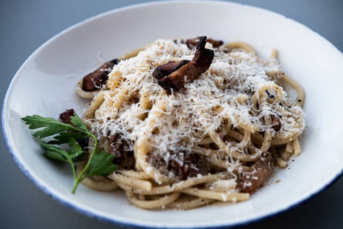 A dish of long, round strands of pasta with grated cheese, hunks of guanciale, and a sprig of parsley.