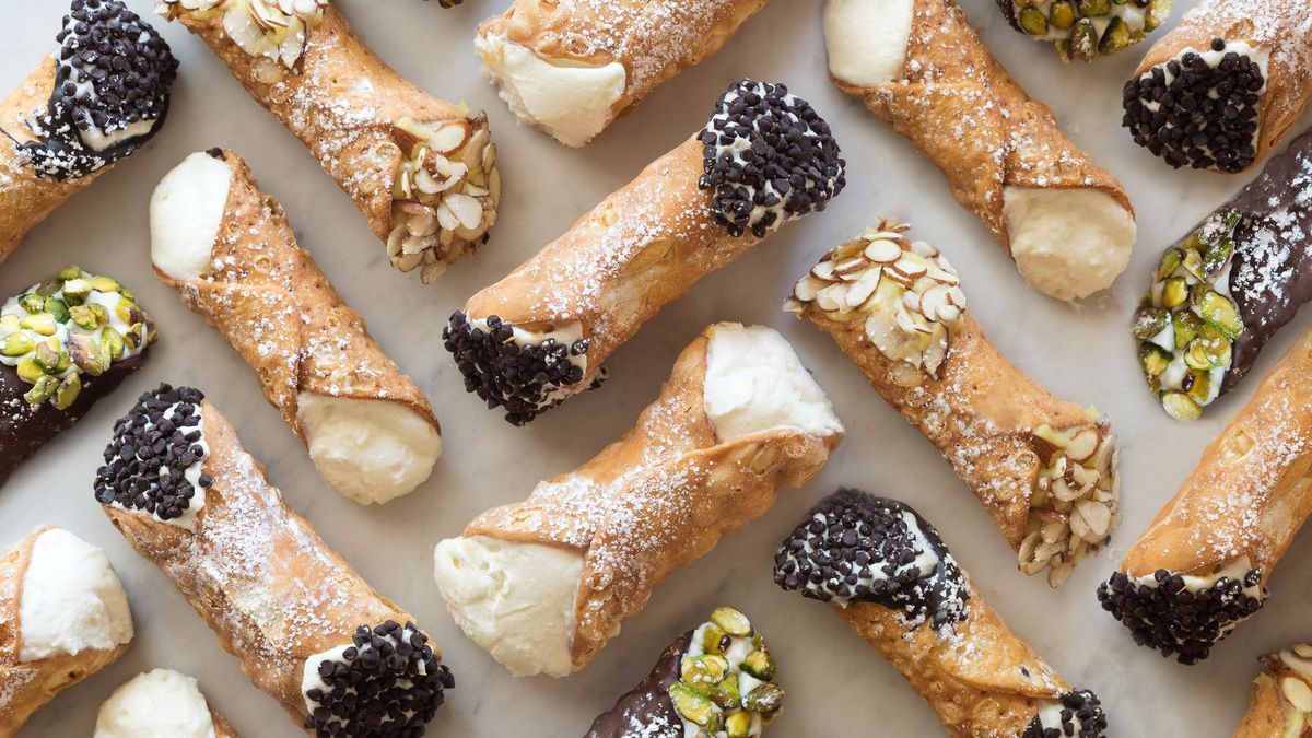 A zig-zag pattern of cannoli from Mike’s Pastry and Modern Pastry, arranged on a white background. Some are garnished with chocolate chips or pistachios.